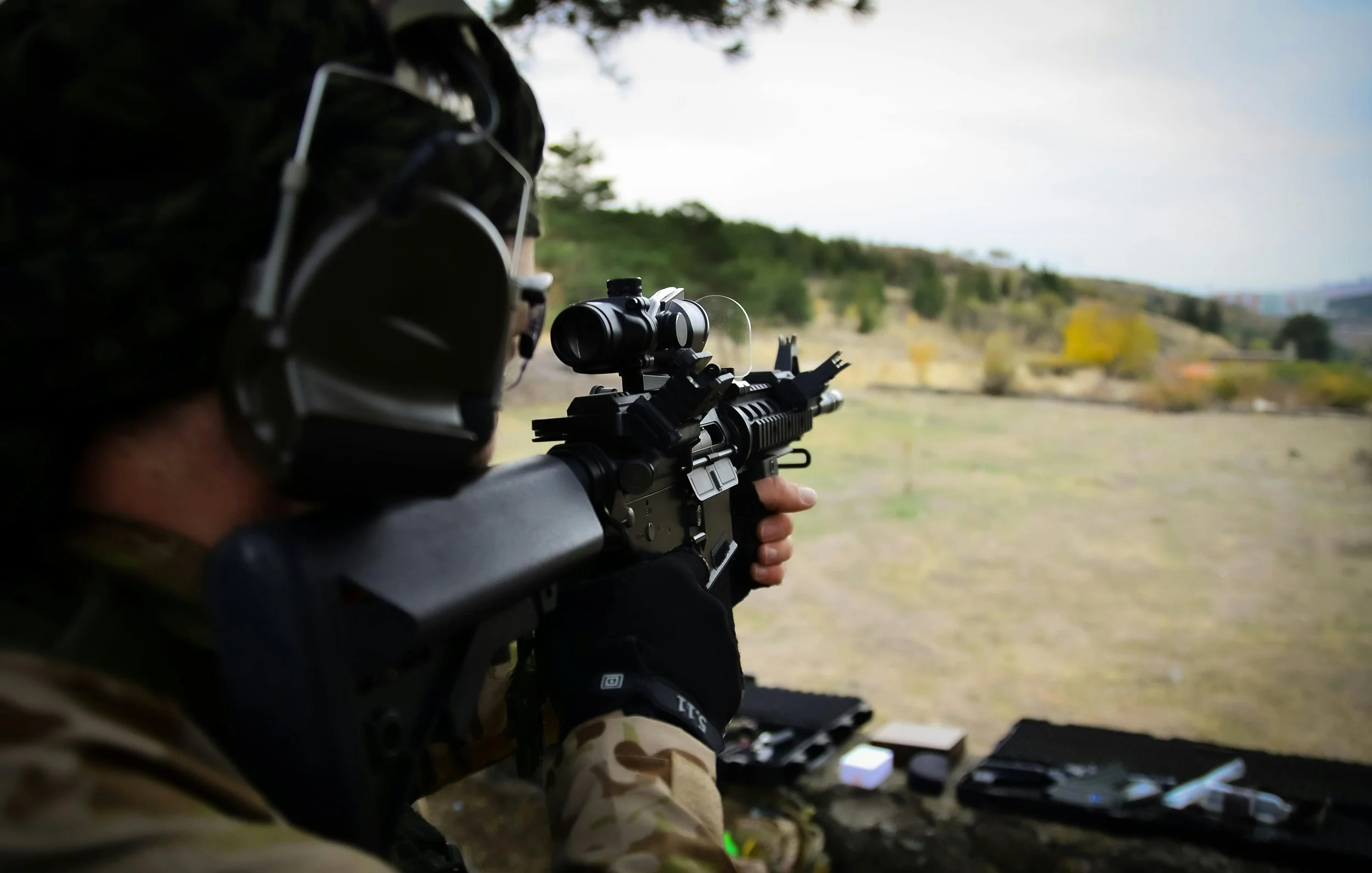 Person wearing camouflage and earmuffs aiming a rifle with a scope at a distant target outdoors.