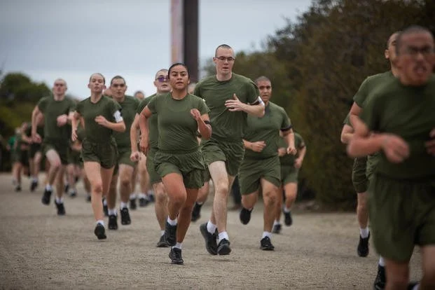 Group of military trainees in green uniforms running outdoors on a dirt path.