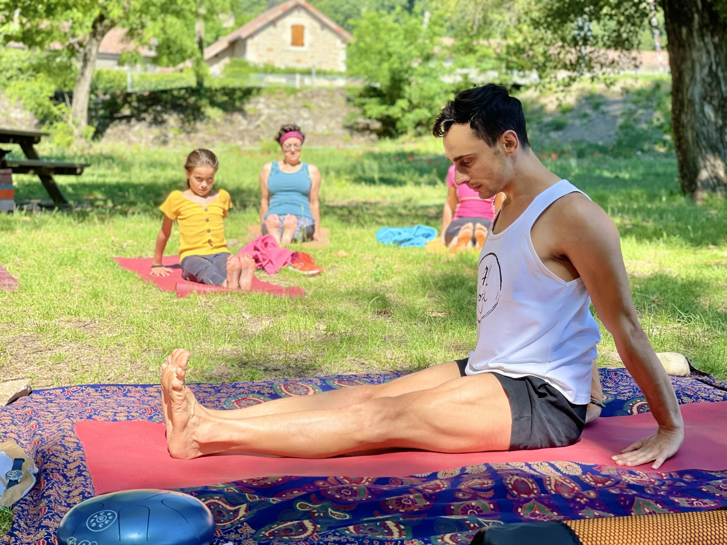 Un jeune homme pratique le yoga en plein air sur un tapis coloré, avec une femme et deux enfants en arrière-plan, dans un parc verdoyant entouré d'arbres et de maisons.