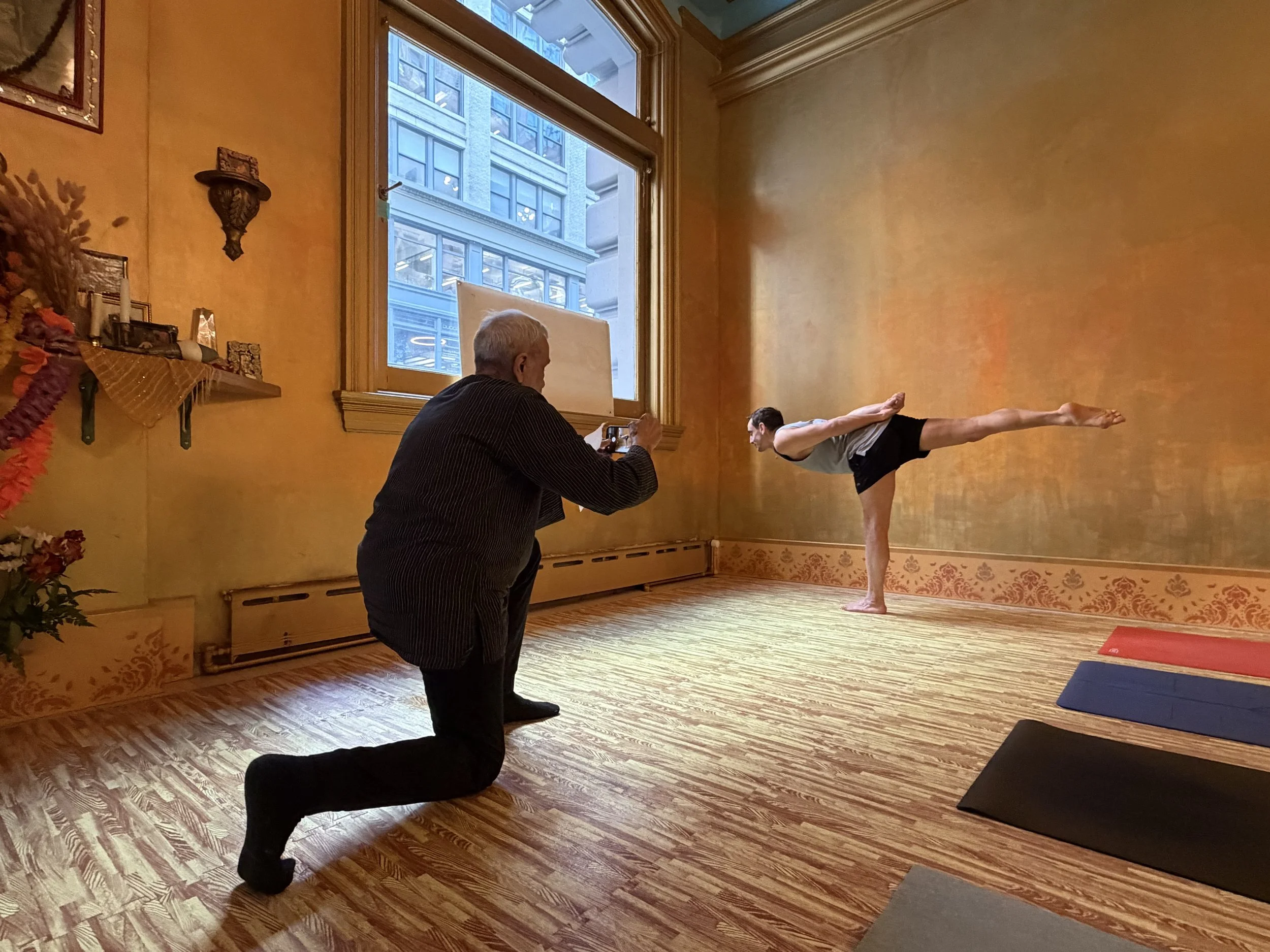 Une personne en position de yoga en équilibre sur une jambe, avec l'autre jambe tendue derrière elle, dans une salle avec murs orange et un grand fenêtre, en train de poser pour une photo prise par une autre personne. 