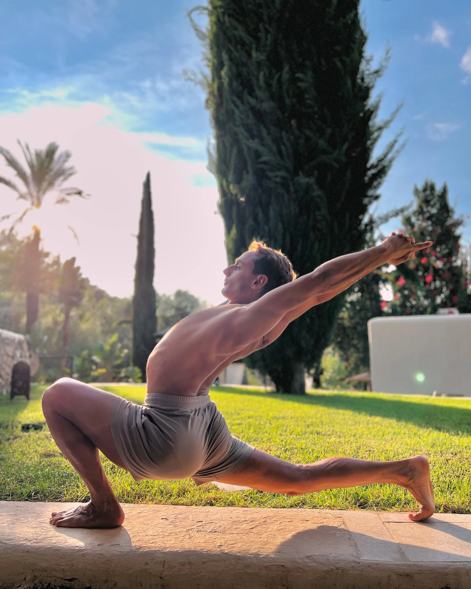 Une personne pratique le yoga en plein air, effectuant une posture de flexibilité sur une plateforme ensoleillée, avec des arbres et un ciel bleu en arrière-plan.