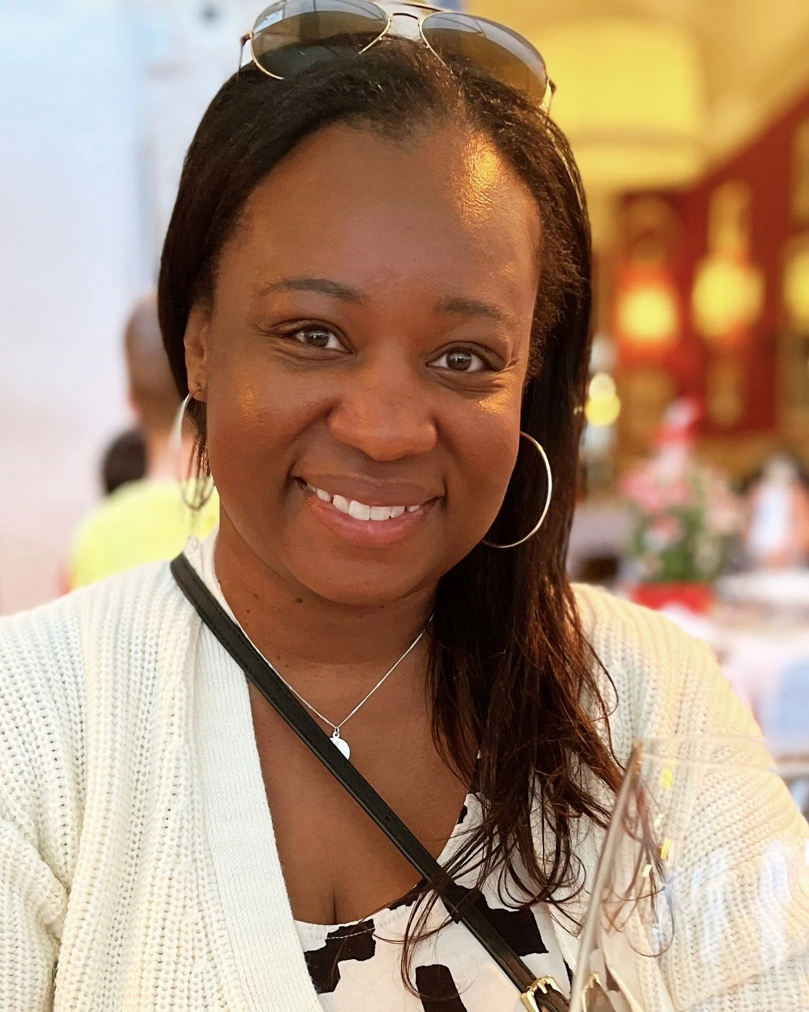 A smiling woman with glasses on her head, wearing hoop earrings and a white sweater, sitting at a table in a warmly lit restaurant or event space.