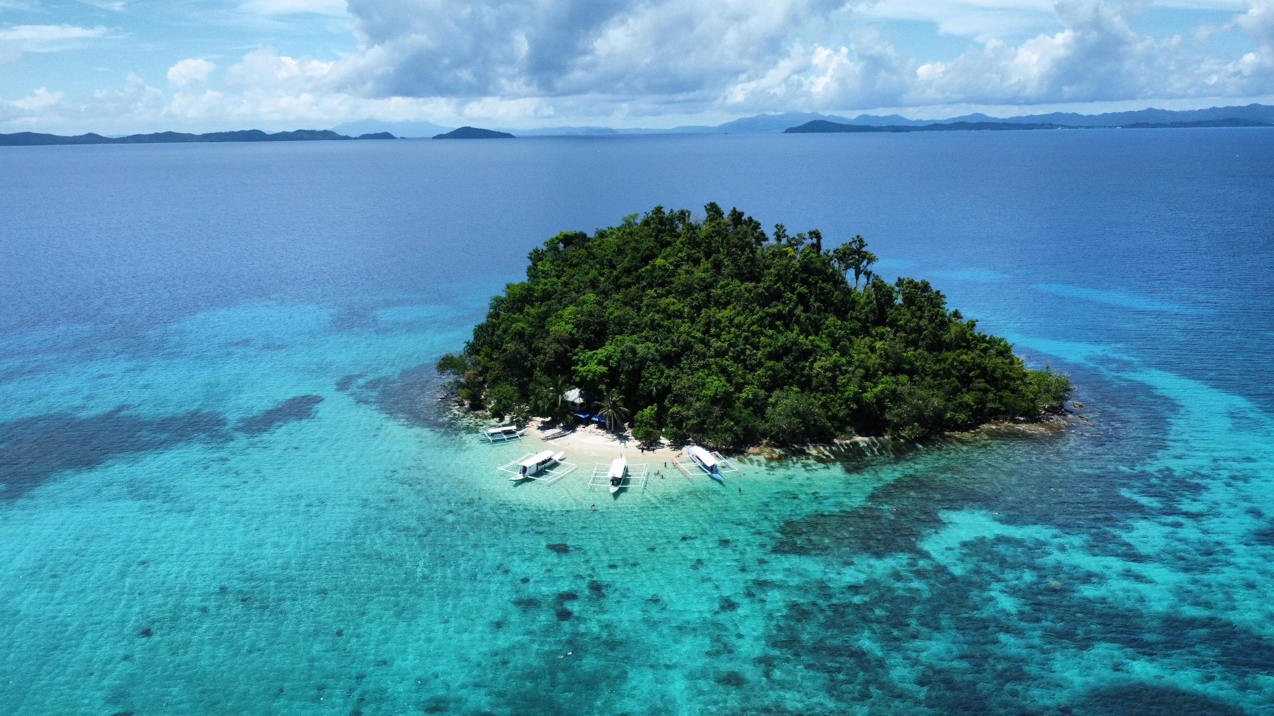 Aerial view of a small tropical island with lush green vegetation, surrounded by turquoise sea and several small boats near the shore.