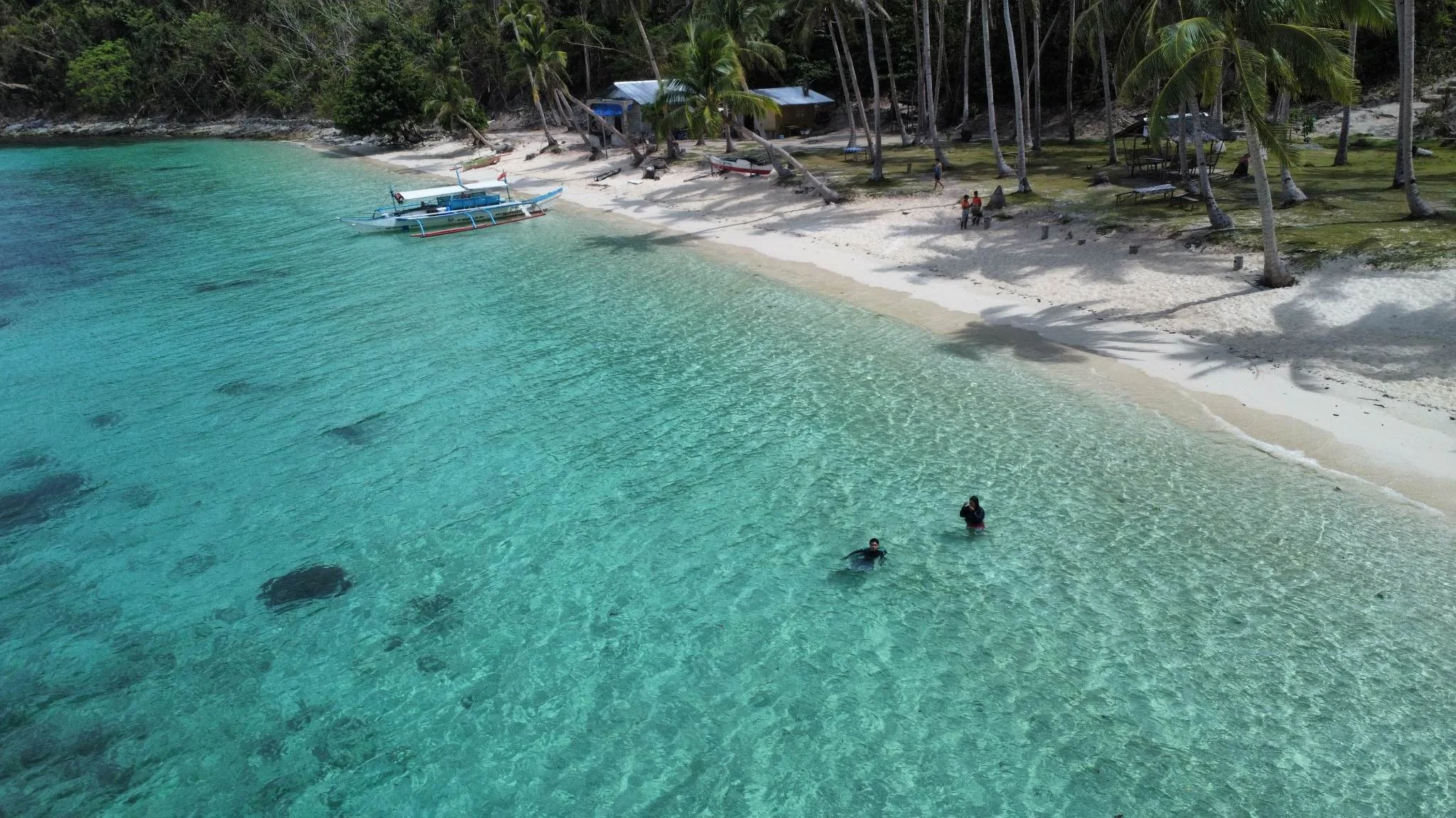 Tropical beach with clear turquoise water, two people swimming, palm trees, white sand, and a small boat anchored near shore.