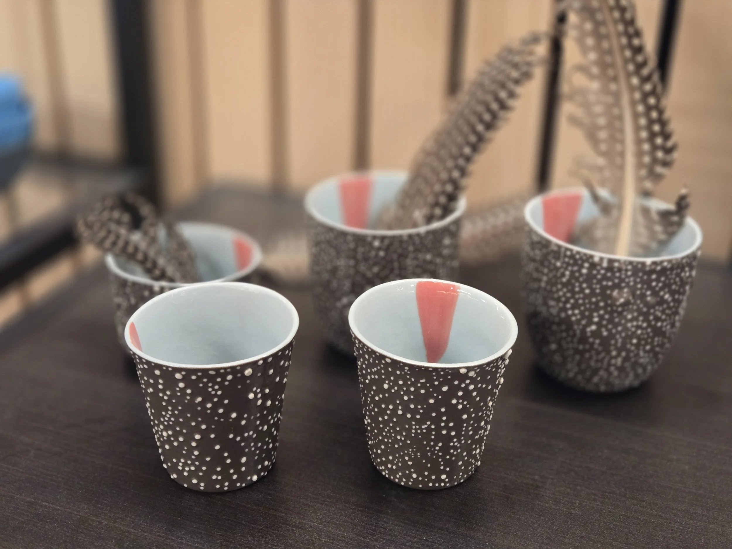 Mehrere Pappbecher mit schwarzen und weißen Punkten, einige enthalten Federn. Die Becher sind auf einem dunklen Tisch platziert.