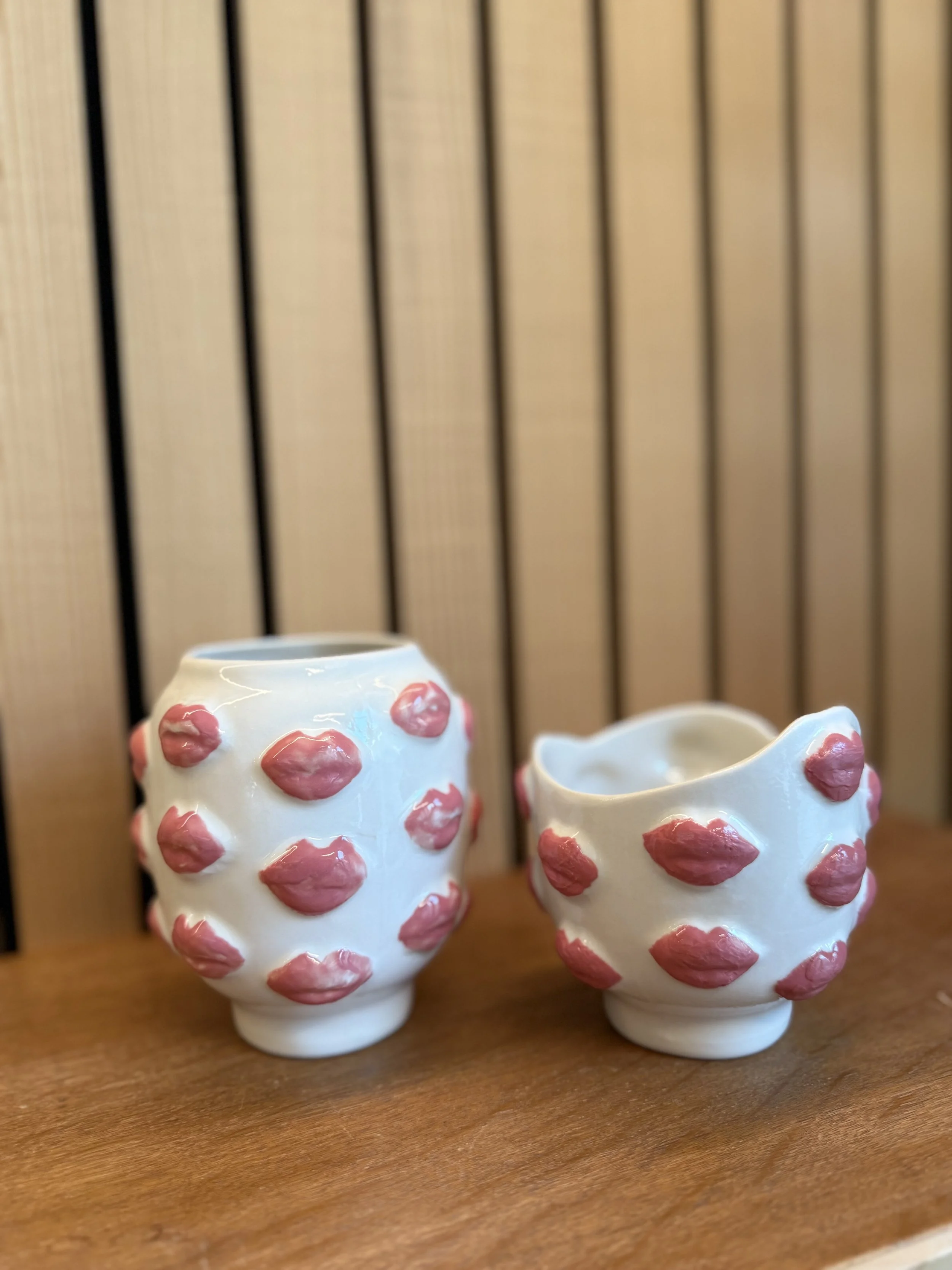 Zwei weiße Keramikvasen mit roten Lippenmustern, eine größere und eine kleinere, stehen auf einem Holztisch vor einer vertikalen Holzwand.