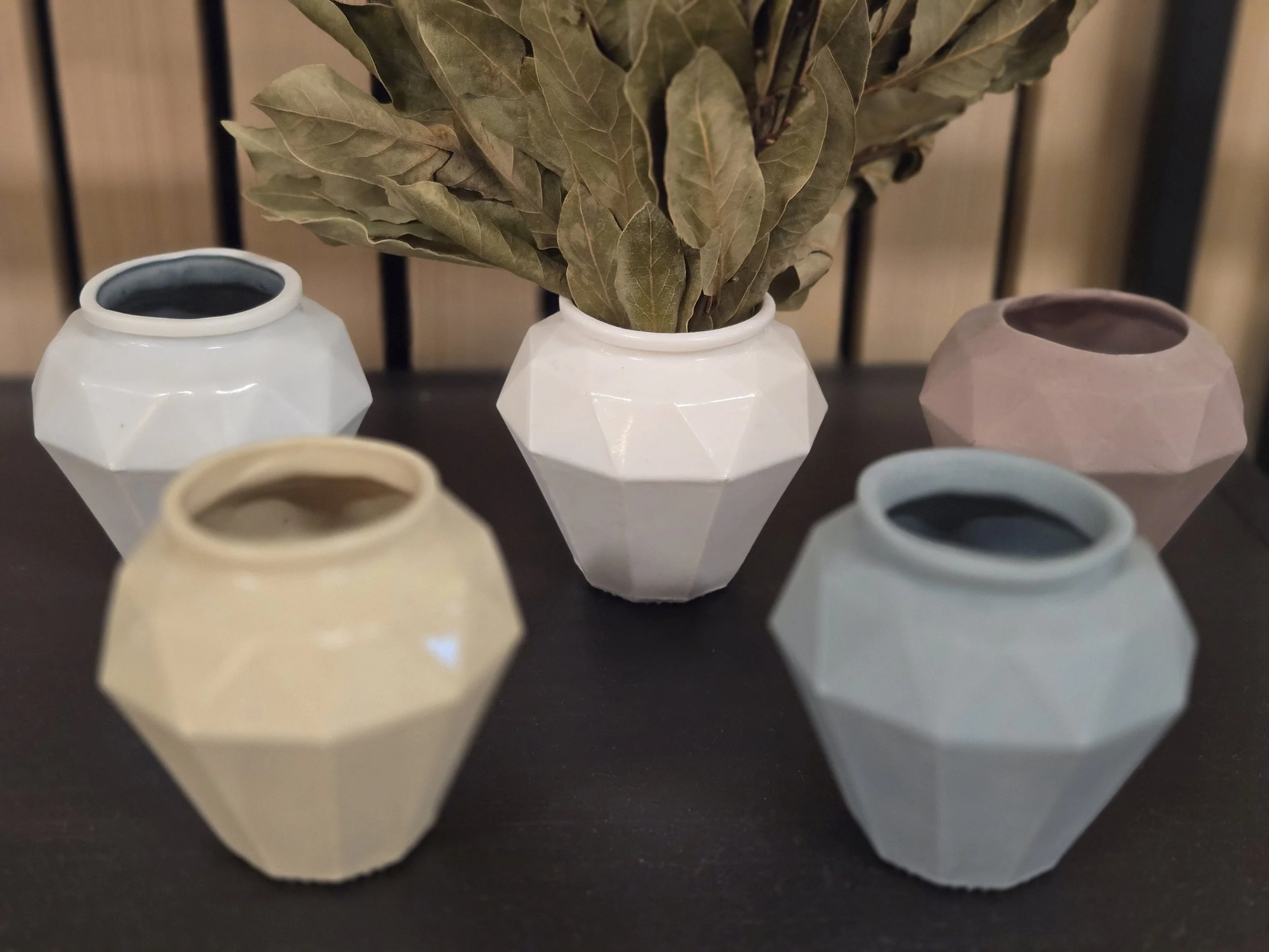 Fünf geometrische Keramikvasen in Pastellfarben, eine mit getrockneten Blättern, auf einem schwarzen Tisch.