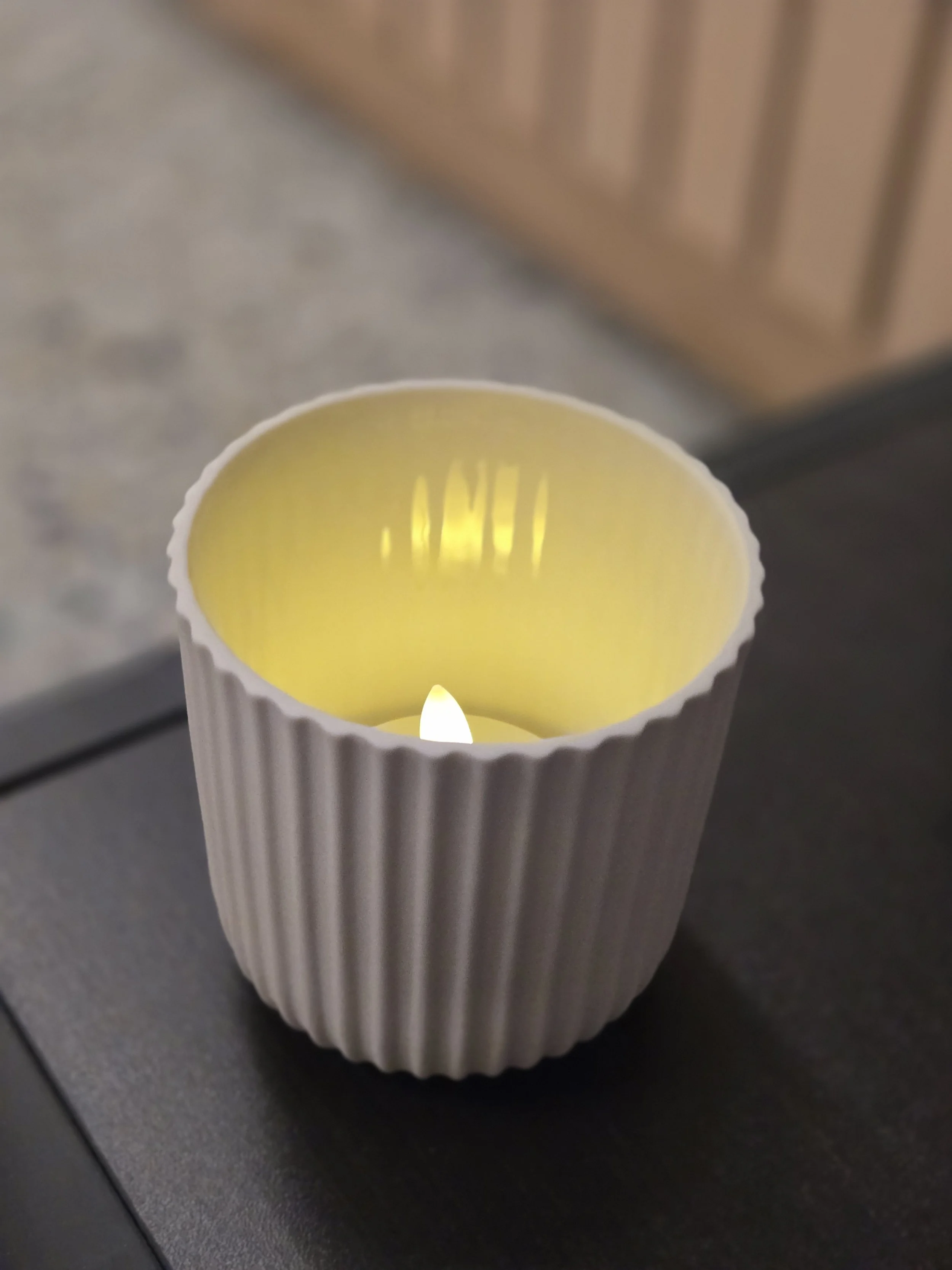 Weiße Kerze in einer vertikalen, gewölbten, gerippten Kerzenglas mit gelber Flamme, auf einer dunklen Oberfläche. Hintergrund aus Holz und grauem Stein.