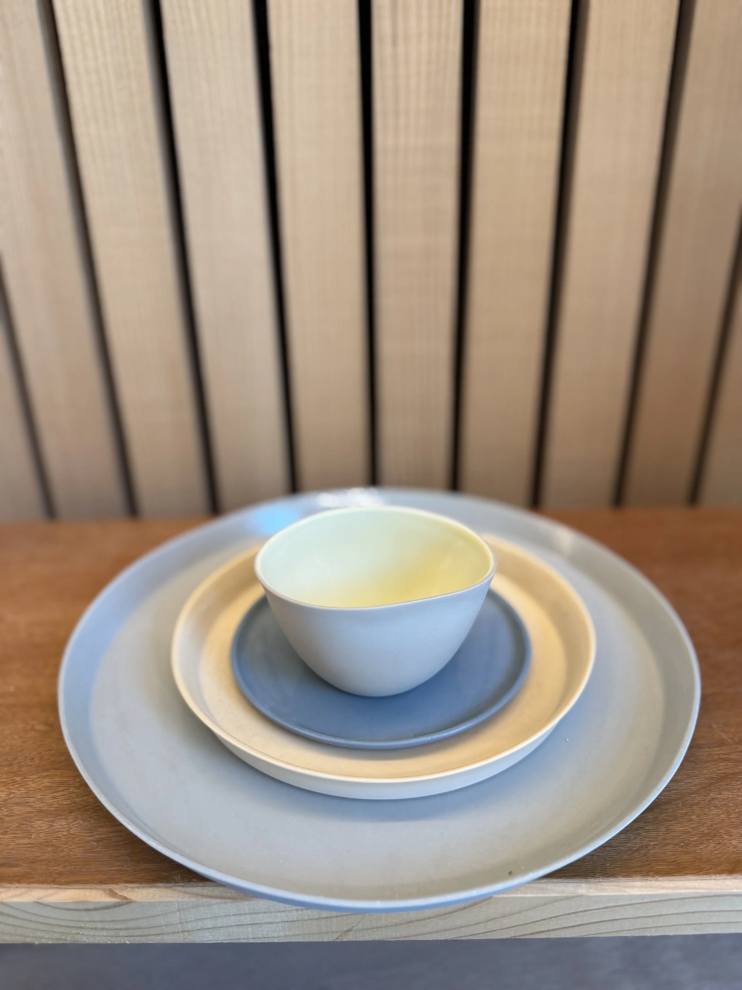 Weiße Teetasse mit einem Suppenlöffel auf einem größeren Teller, auf einem Holztablett, vor einer Holzverkleidung im Hintergrund.