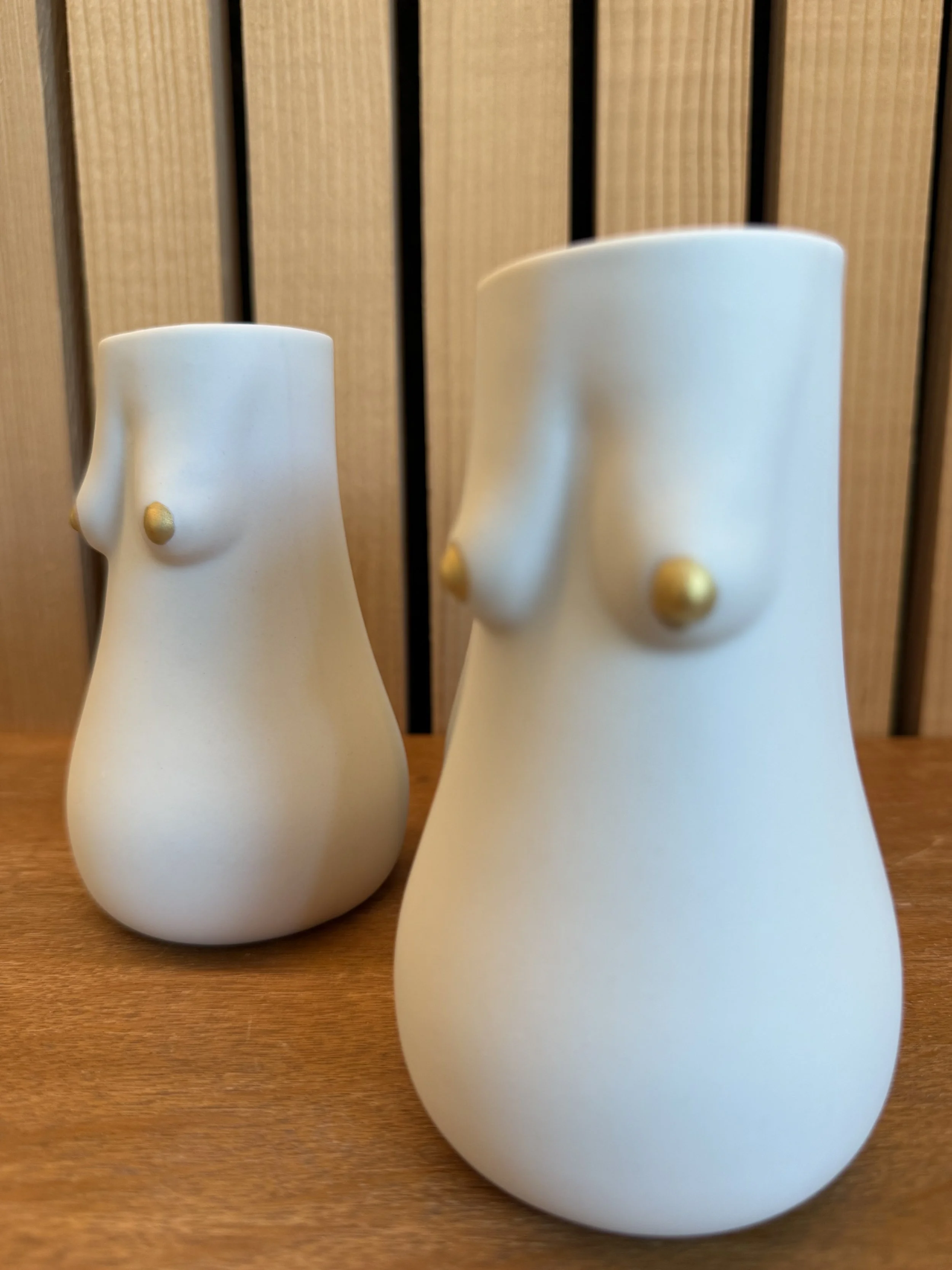 Zwei Schuhe in Form von menschlichen weiblichen Figuren mit stilisierten Brüsten, weiß mit goldfarbenen Brustwarzen, auf einem Holztisch vor Holzlattenwand.