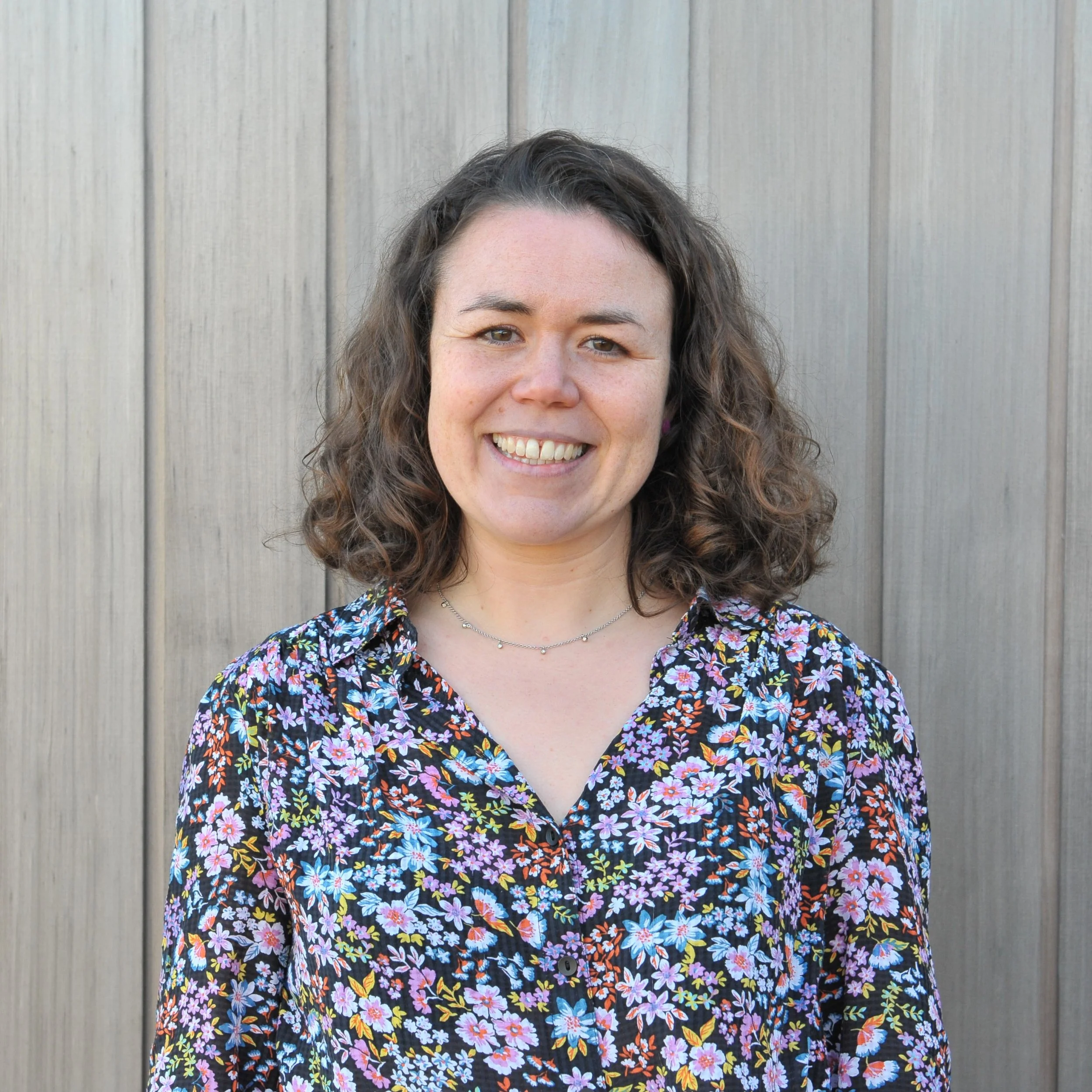 A woman with shoulder-length curly brown hair, smiling, wearing a black jacket over a colourful floral dress, standing in front of a building.