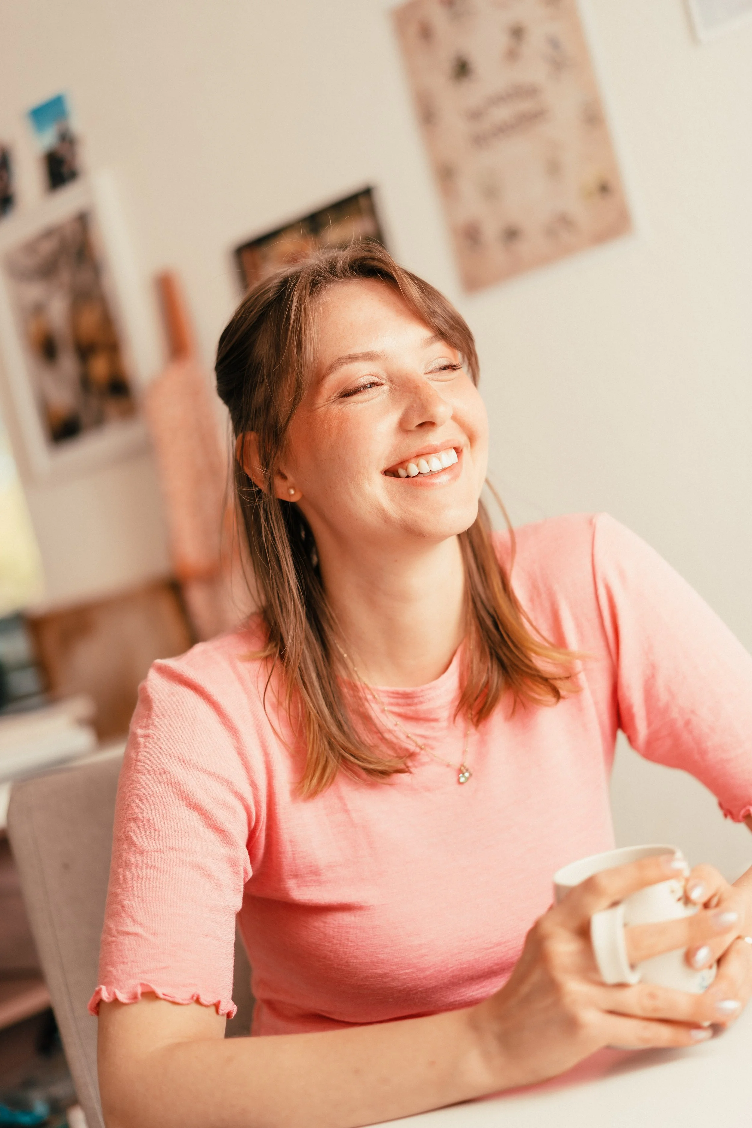 Freudige junge Frau mit braunem Haar in rosa T-Shirt, die einen weißen Kaffeetasse hält, lächelnd im Raum mit Fotos und Postern an der Wand.