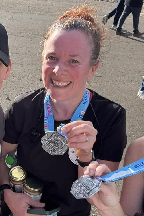 Woman smiling and holding a medal at a race event, wearing a black shirt and a blue race bib, with others in the background.