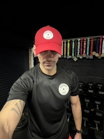 Man wearing a red cap and black athletic shirt in a gym with medals hanging on a rack in the background.