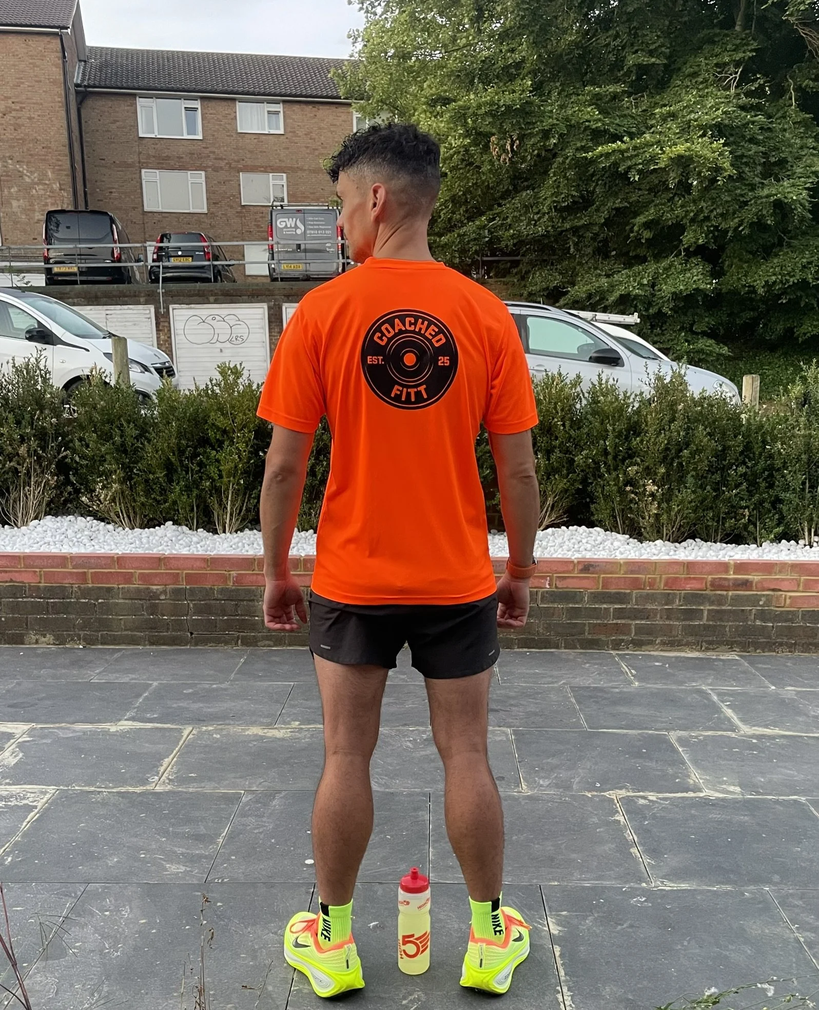 A man with short hair, wearing a bright orange athletic shirt with a round logo on the back that reads 'COACHED FITT' and 'EST. 25,' black shorts, neon yellow Nike running shoes, and neon yellow socks with red accents, standing outdoors on a paved surface with his back to the camera. There is a water bottle in front of him.