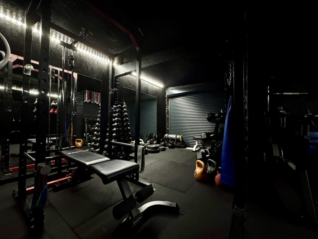 A dimly lit gym with various workout equipment including a weight bench, dumbbells, kettlebells, and resistance bands.