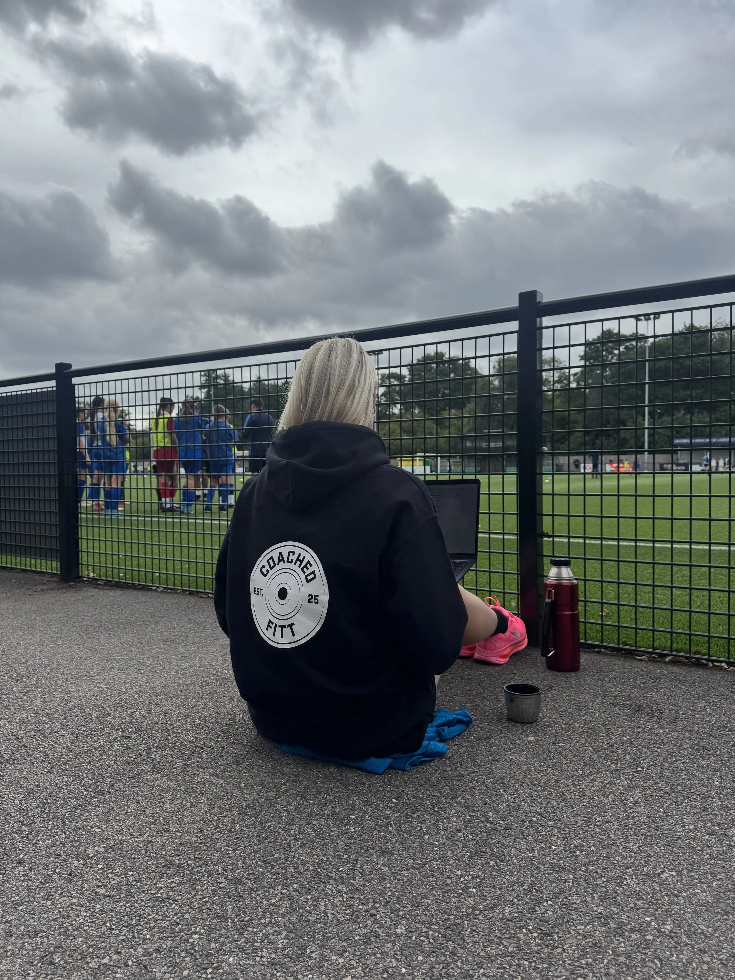 A person with blonde hair sitting on the ground outside a soccer field, watching a game. They are wearing a black hoodie with a logo on the back that says 'Coached Fitt'. They have a laptop, pink sneakers, a reusable water bottle, a tumbler, and a small container nearby. The soccer game is underway, with players in blue and red uniforms, and the sky is overcast.