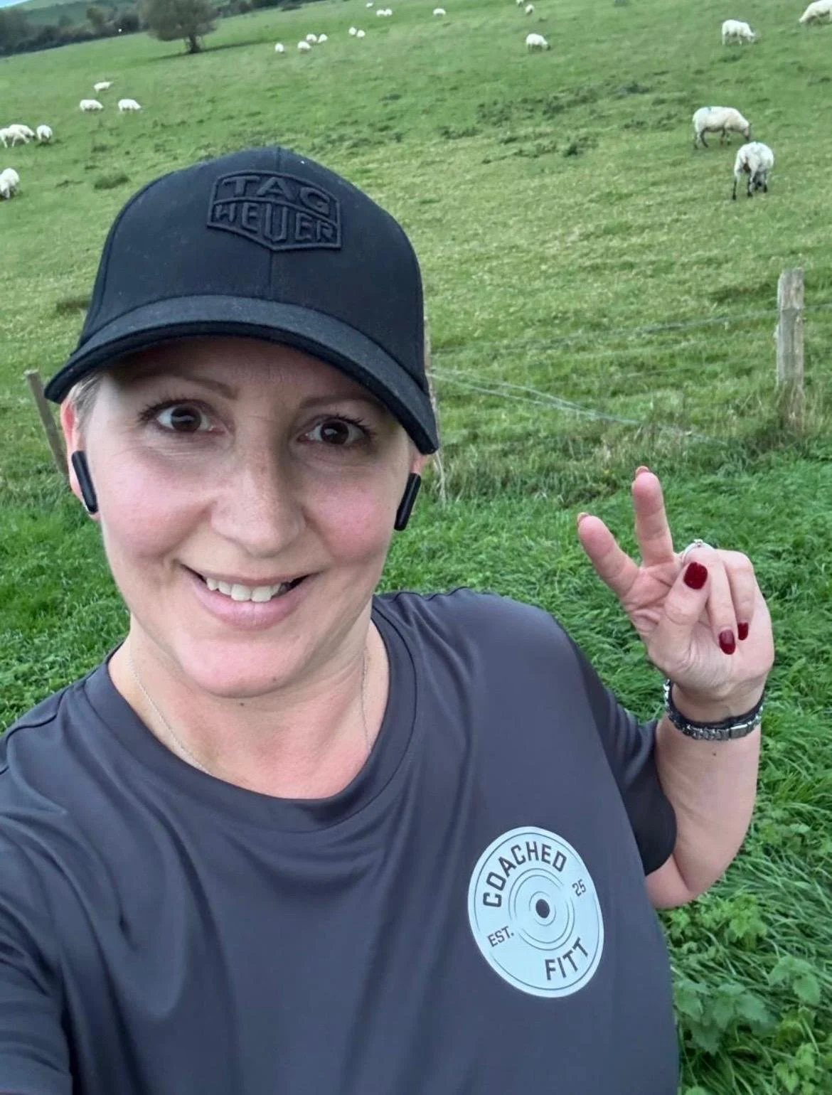 A woman smiling and taking a selfie in a green field with sheep grazing in the background. She is wearing a black Tag Heuer cap, a dark athletic shirt with a