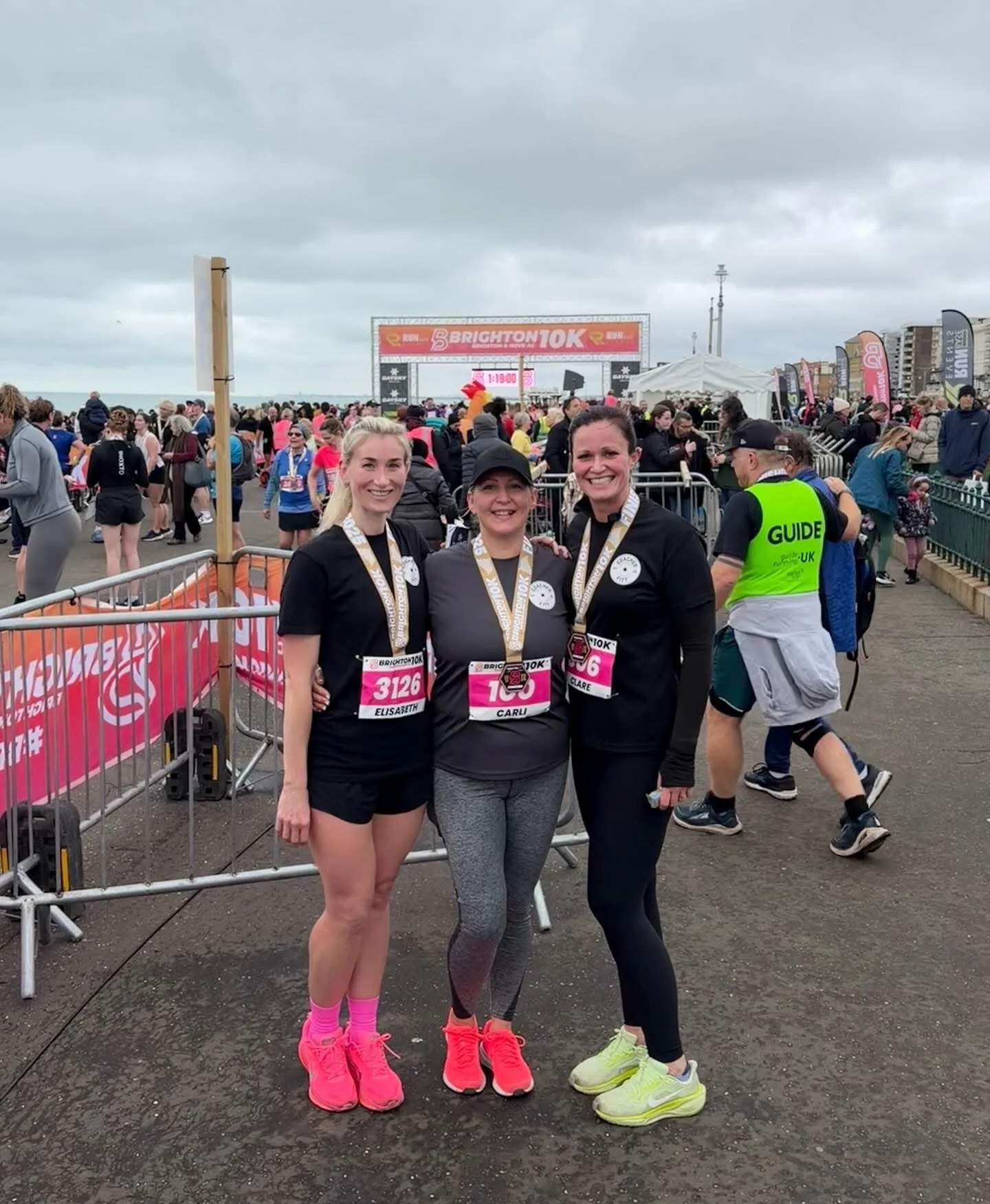 Three women wearing medals and race bibs at the Brighton 10K race event, standing in front of a crowd and finish line, with gray cloudy sky overhead.