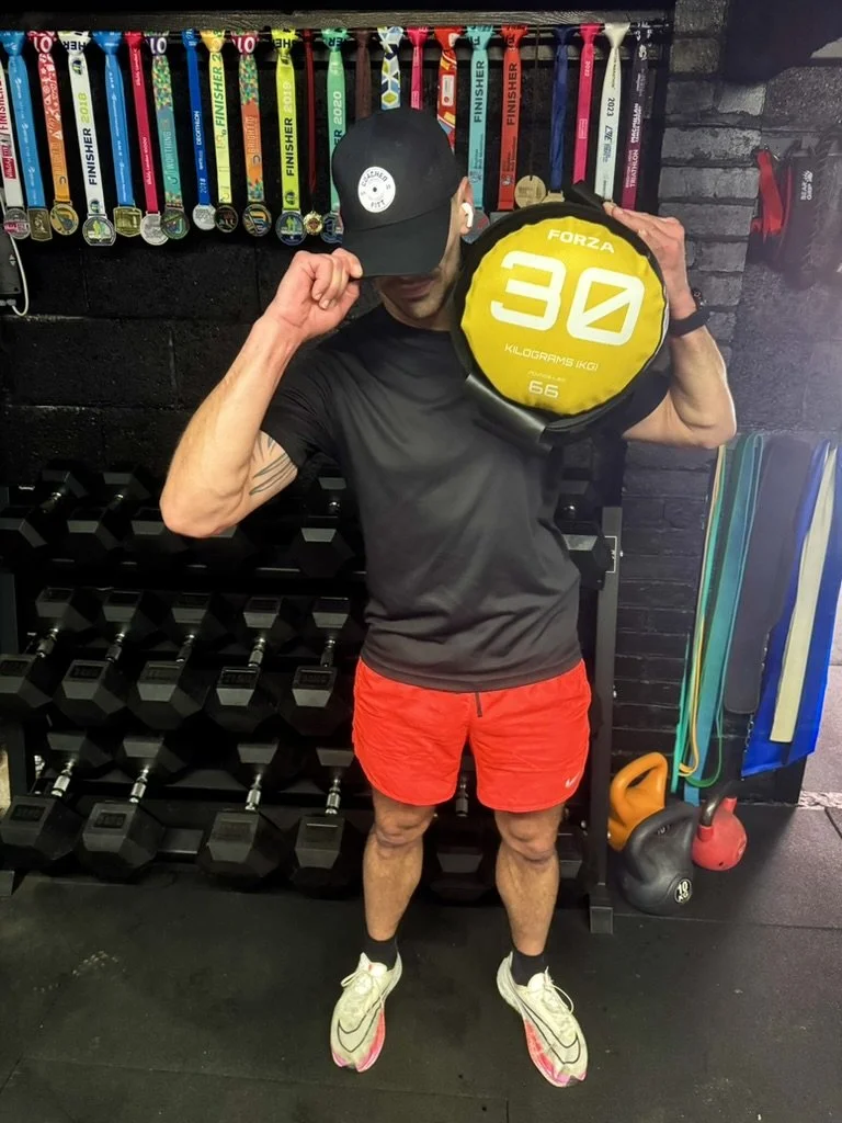 Man holding a Forza 30-kilogram weight plate in a gym, wearing a black cap, black shirt, red shorts, and running shoes, standing in front of a wall with medals and fitness equipment.