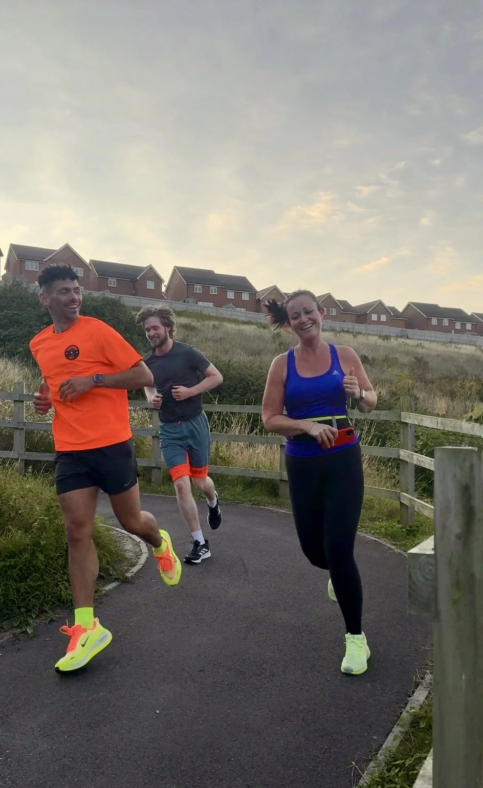Three people running outdoors on a paved trail, with houses in the background and a cloudy sky overhead. Two men and one woman are smiling and appear to be enjoying their run.