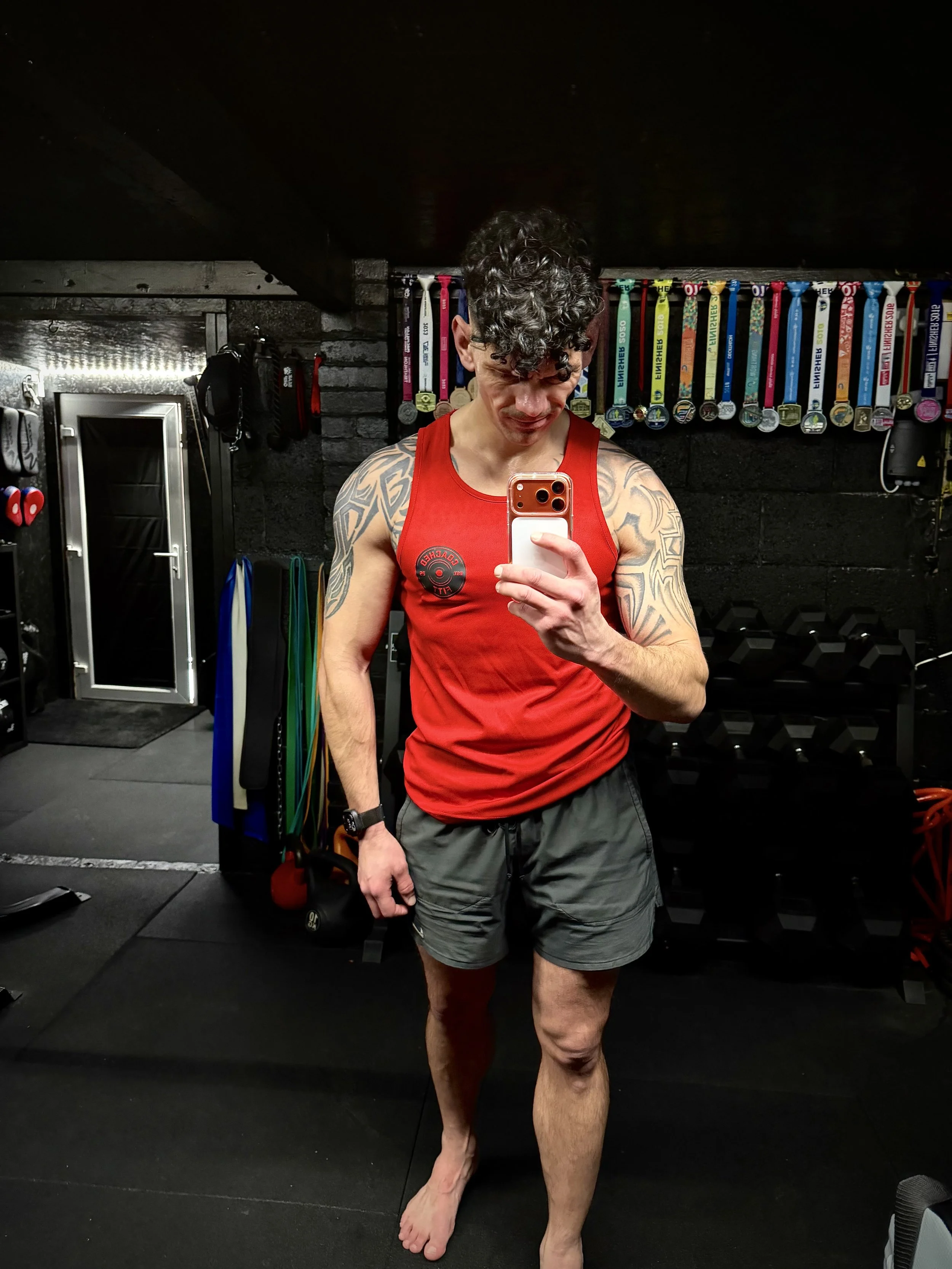 A young man taking a mirror selfie in a gym. He has curly dark hair, tattoos on his arms, and is wearing a red sleeveless shirt and gray shorts. Medals are hanging on the wall behind him.