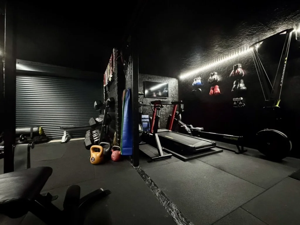 A home gym with boxing gloves on the wall, a treadmill, a rowing machine, kettlebells, dumbbells, medicine balls, and a television, all in a dark room with black walls and ceiling.