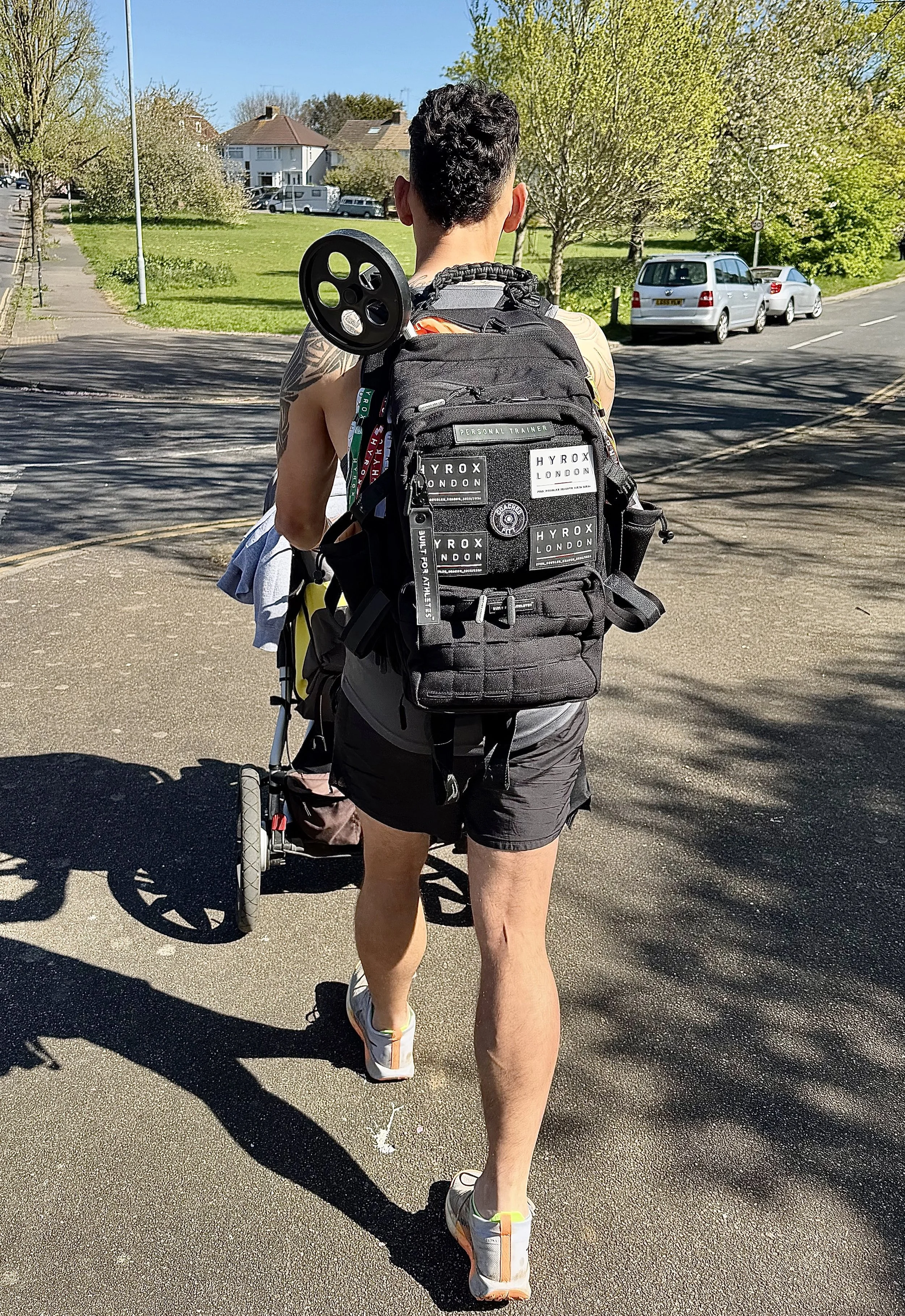 A man with tattoos and wearing sneakers is walking on a sidewalk, pulling a child in a stroller. The man is carrying a backpack with patches. There are trees, parked cars, and houses in the background on a sunny day.