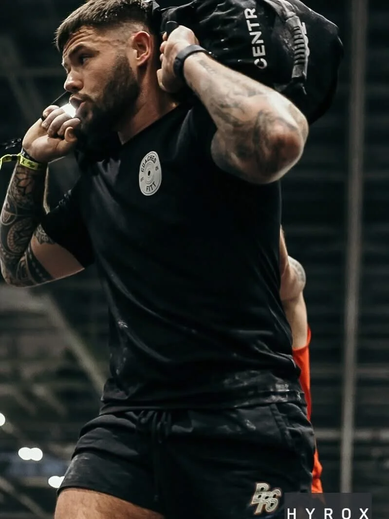 A man with tattoos on his arms is carrying a black gym bag on his shoulder in a gym setting, wearing a black shirt and shorts.