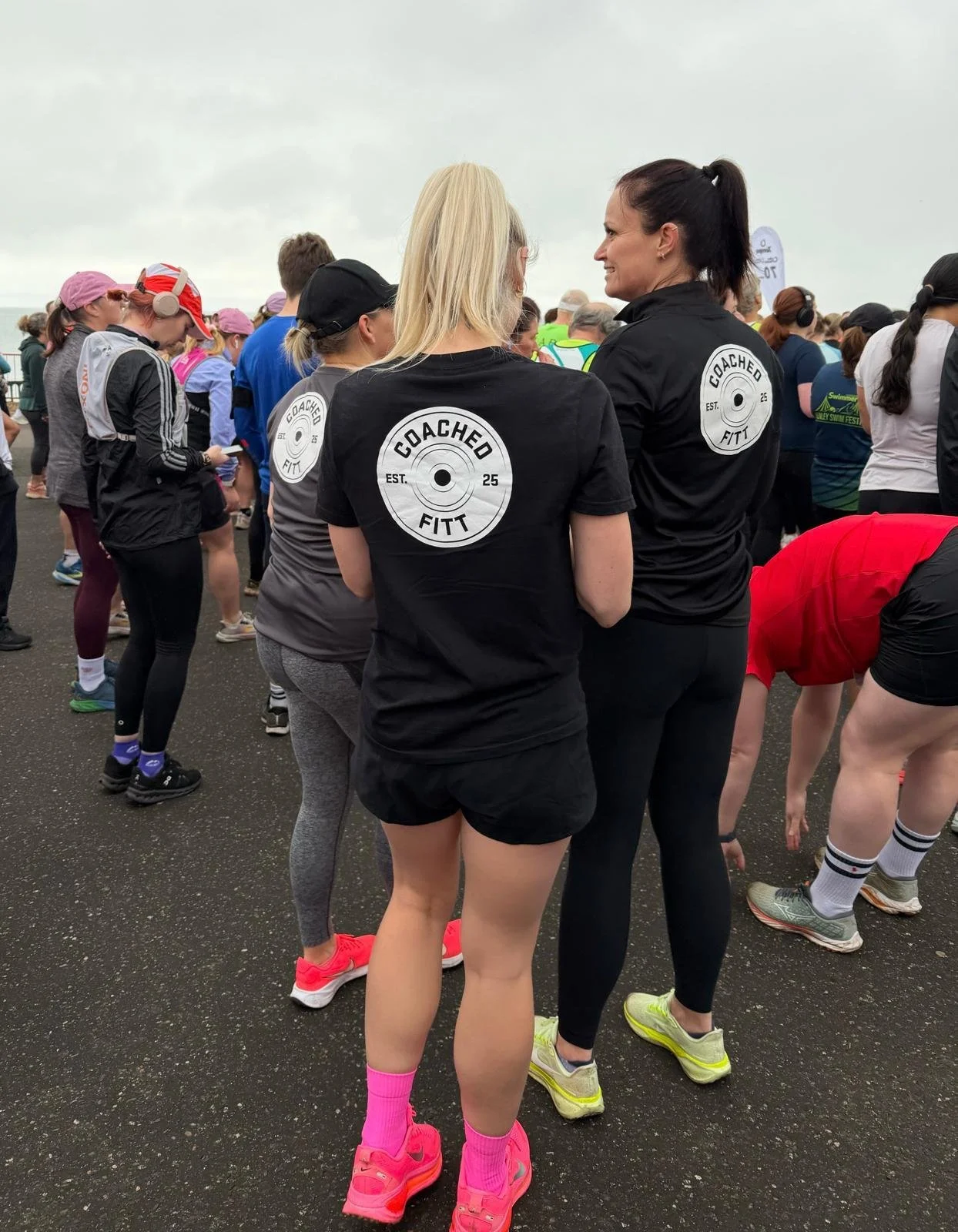 Two women wearing black shirts with 'Coached FITT' logos on their backs, standing among a crowd of people in athletic attire, at an outdoor event on a cloudy day.