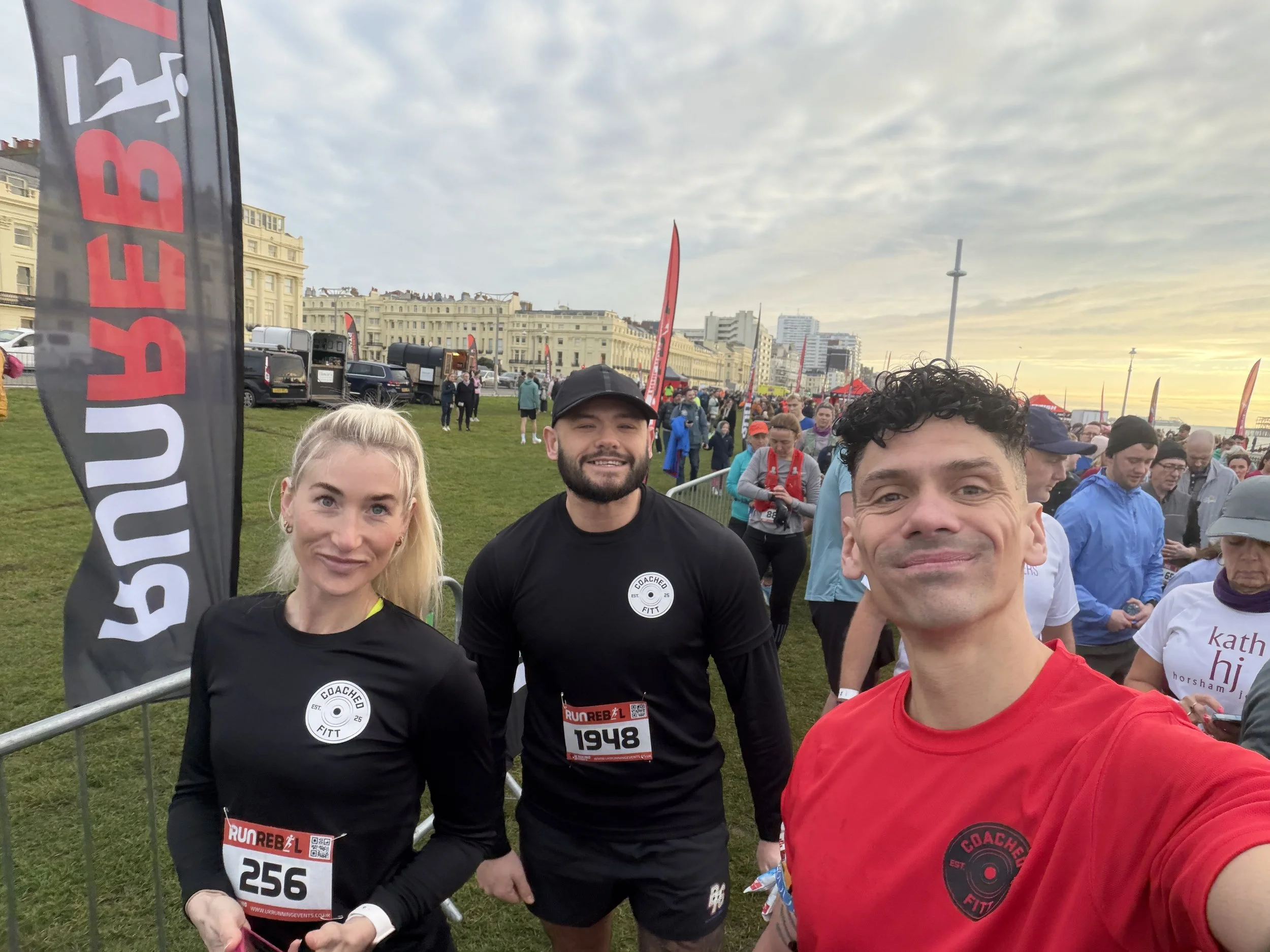 Three individuals taking a selfie at a marathon event, with many other runners and event banners in the background during sunrise or sunset.