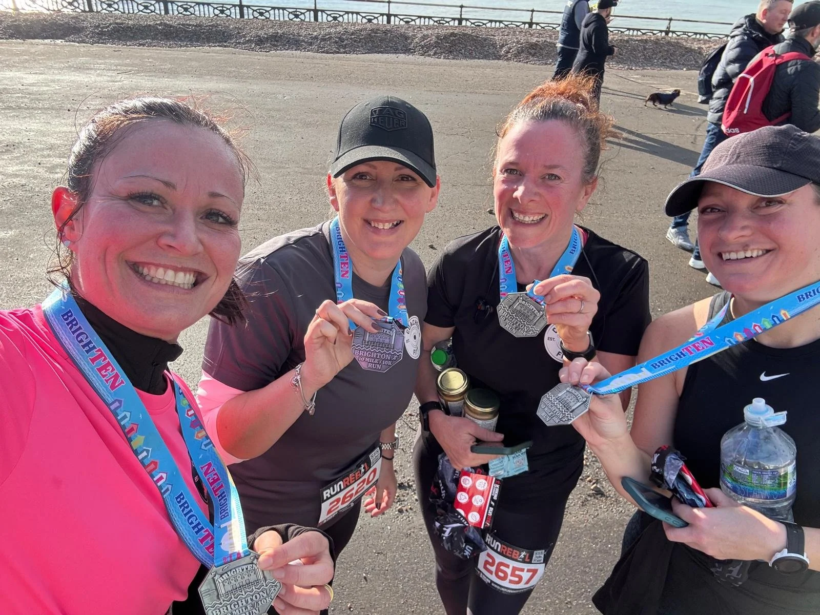 Four women smiling and showing medals after a race, holding water bottles and race bibs, with other runners in the background.
