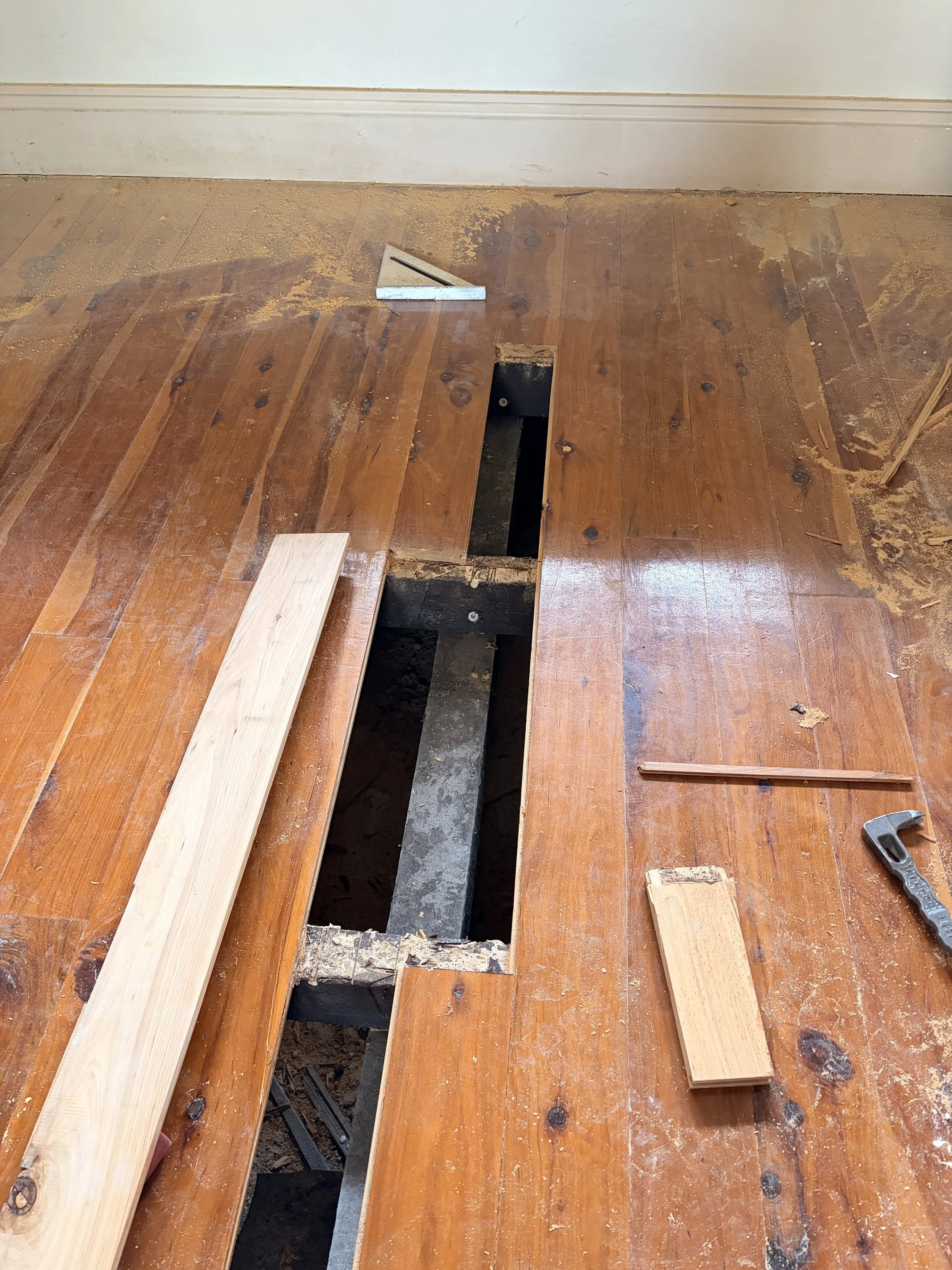 View of a wooden floor under renovation with a hole, a carpenter square, wood pieces, and construction tools scattered around.