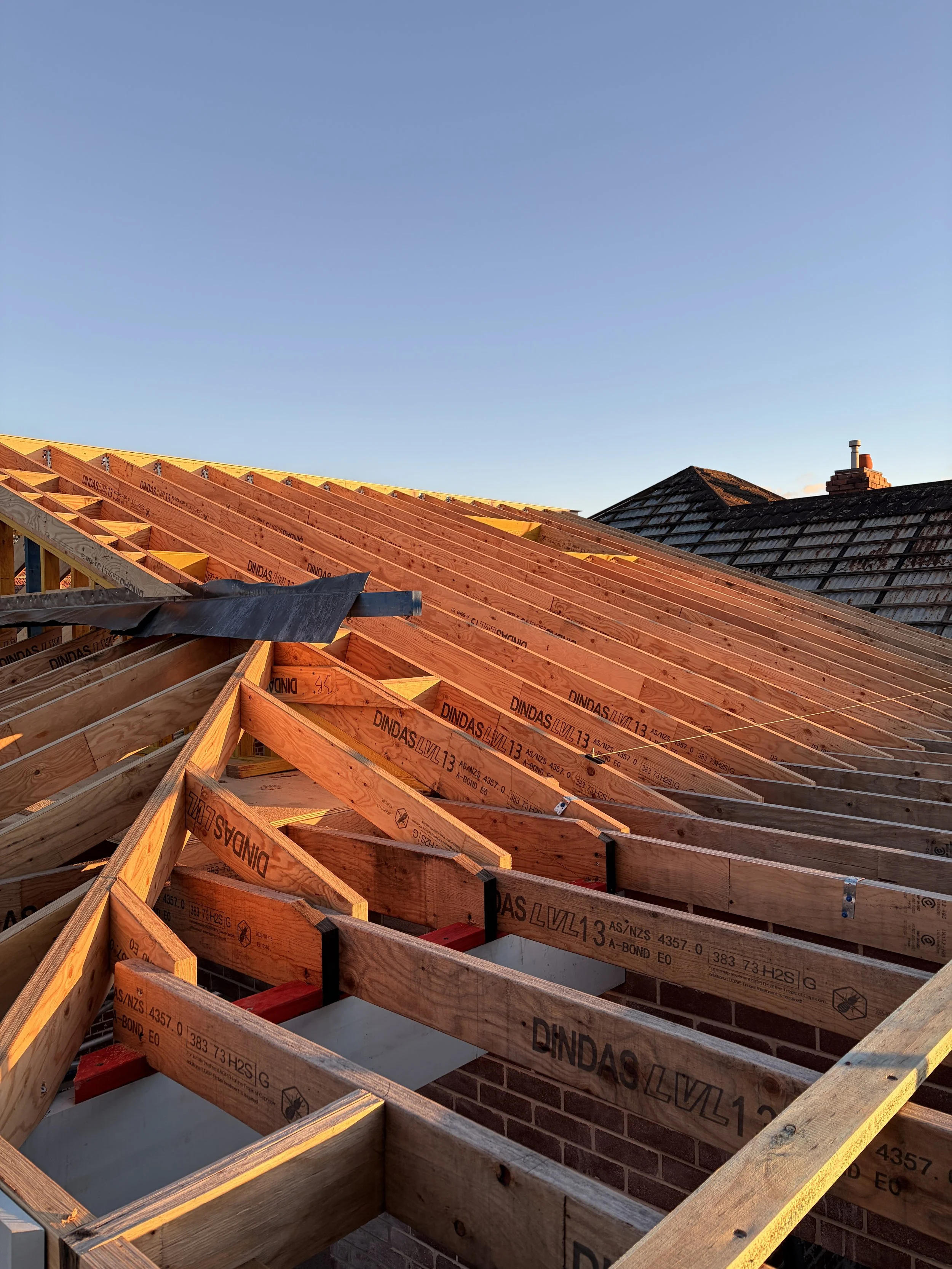 A house roof under construction with wooden framing and a section of existing brick wall, against a clear blue sky.
