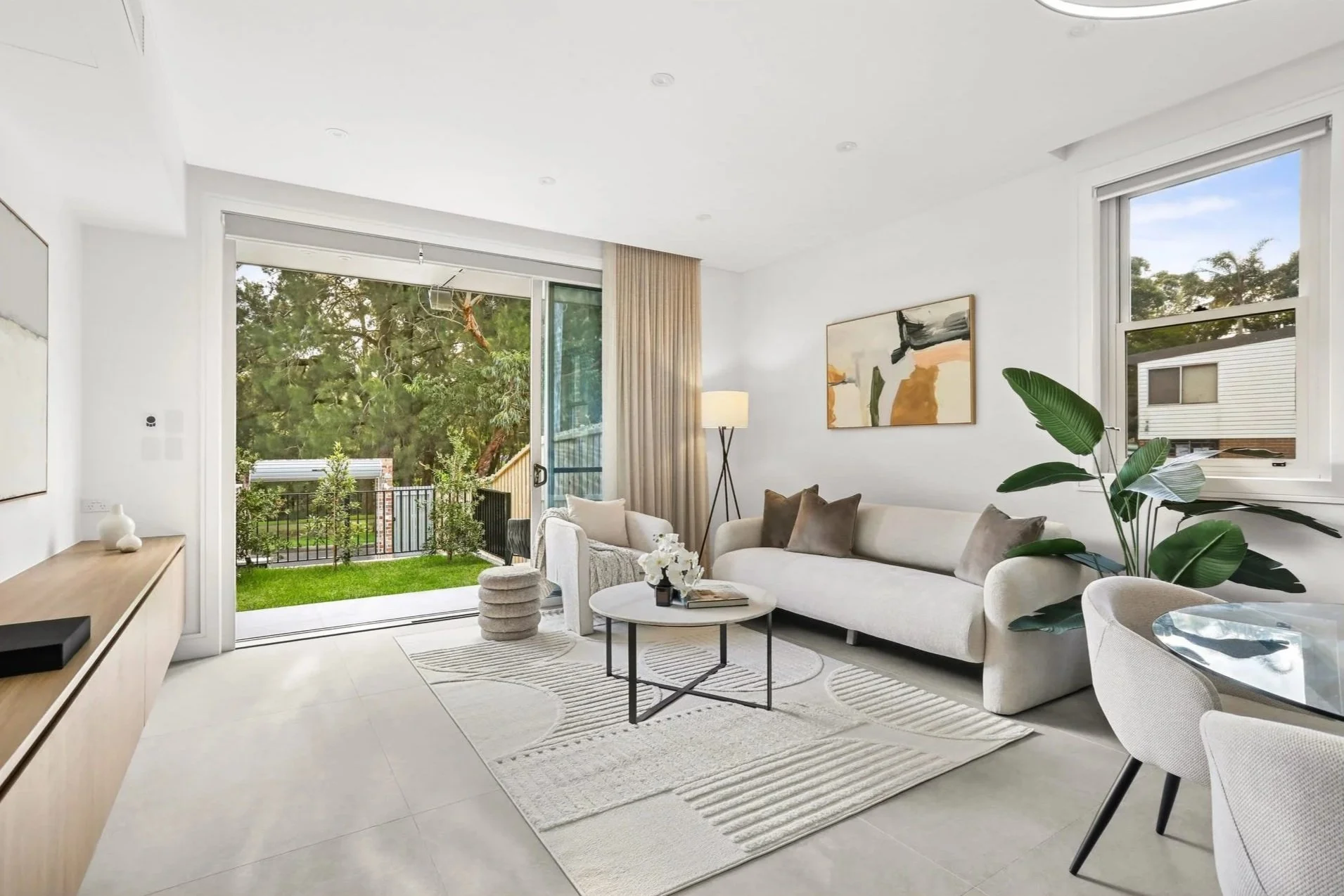 Bright living room with white walls and furniture, large windows showing greenery outside, decorative art on wall, potted plant, and modern furnishings.