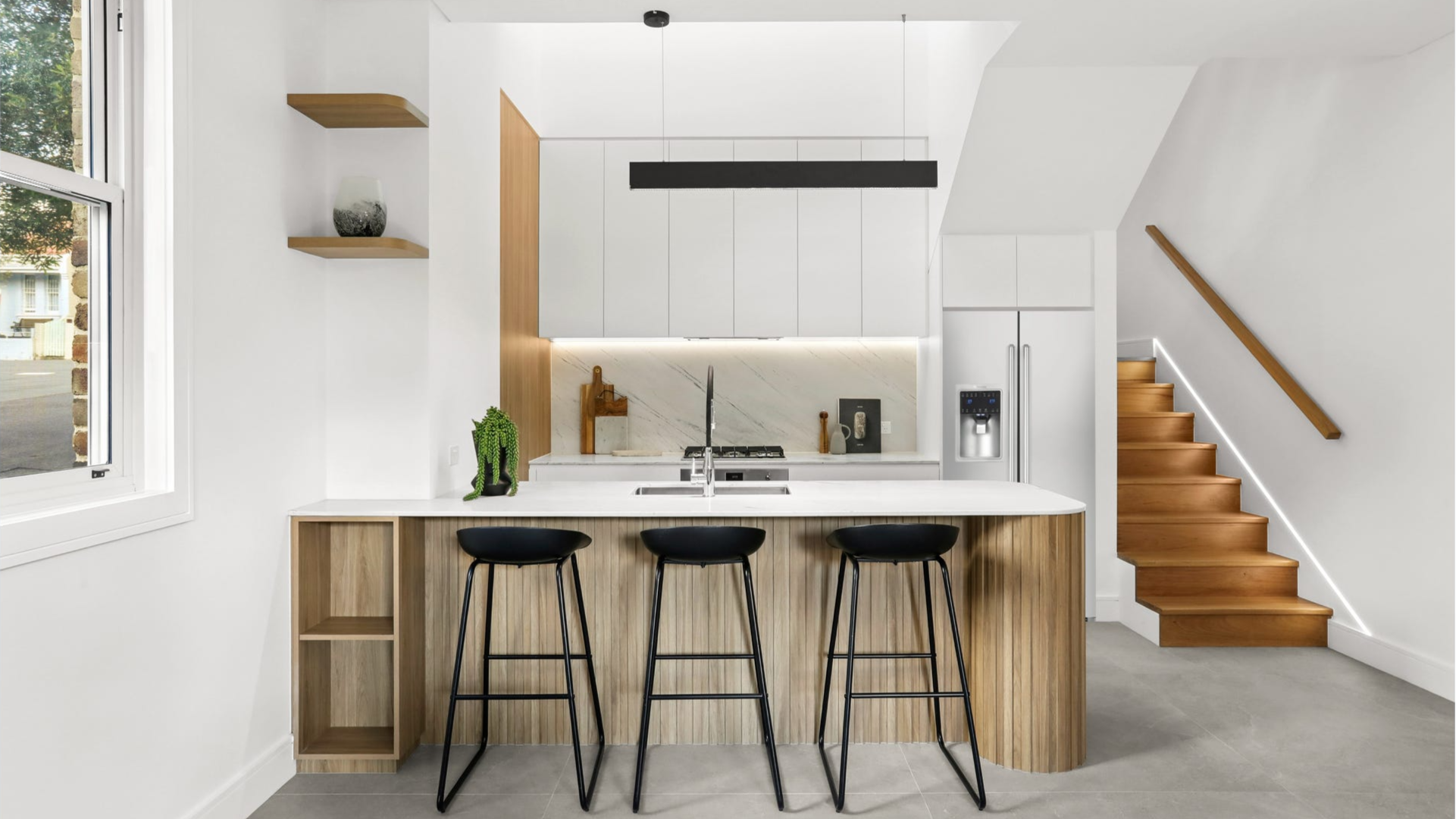Modern kitchen with white cabinets, wooden accents, black stools, and a staircase with wooden steps and LED lighting.