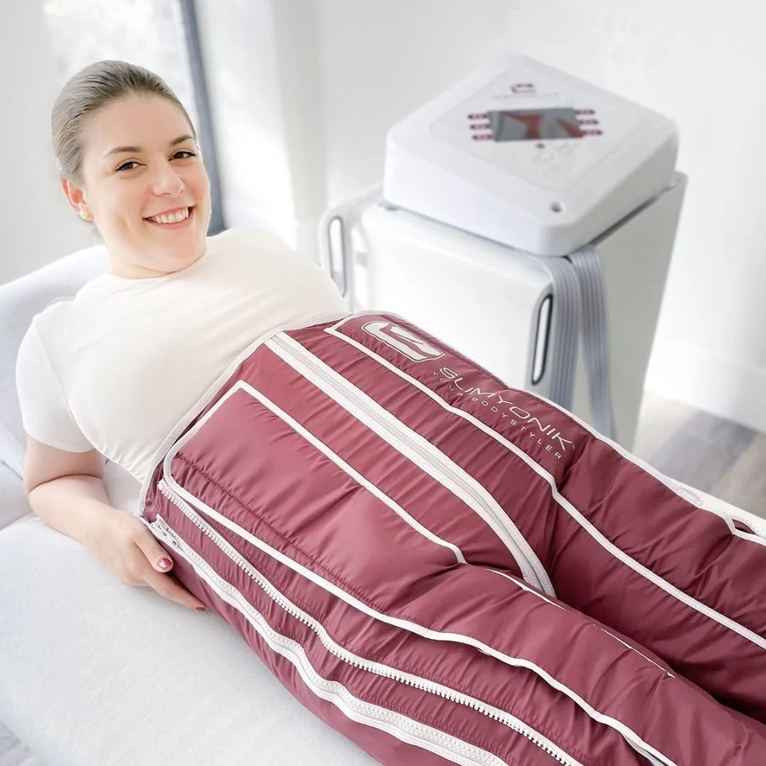 Eine lachende Frau liegt in einer Liege, in der Slimyonik Hose für Lymphdrainagen, während eine medizinische Maschine im Hintergrund steht.