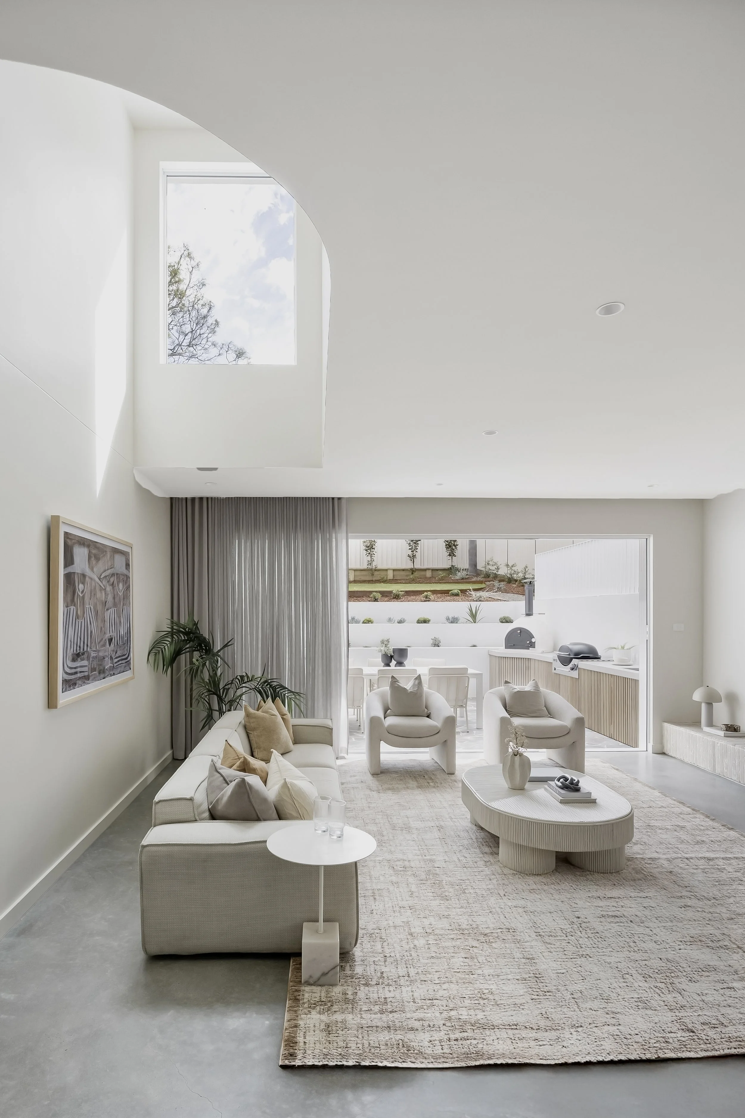 Bright, modern living room with white furniture, neutral-colored pillows, a large area rug, and a view of a patio and garden outside.