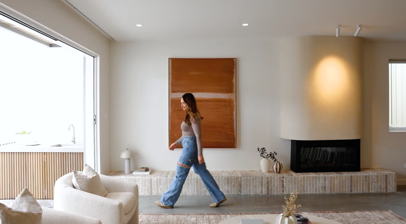 Interior of a modern living room with a woman walking past a fireplace, large window, and minimalist décor.