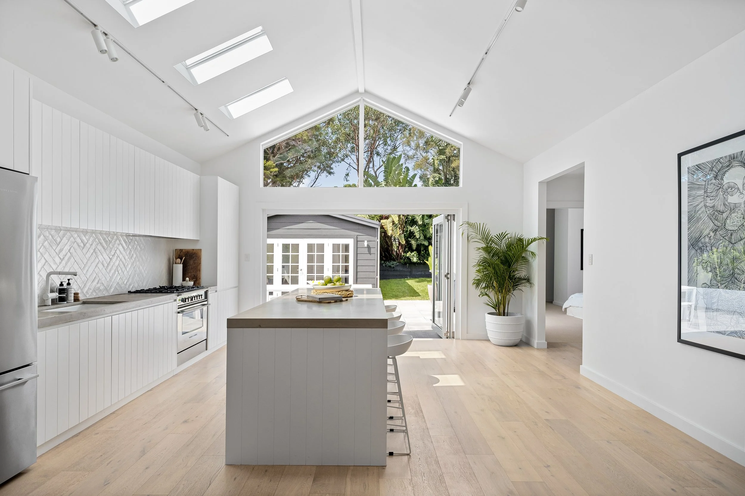 Modern white kitchen with a large island, stainless steel refrigerator, skylights, and open double doors leading to a backyard with a shed and lush greenery.