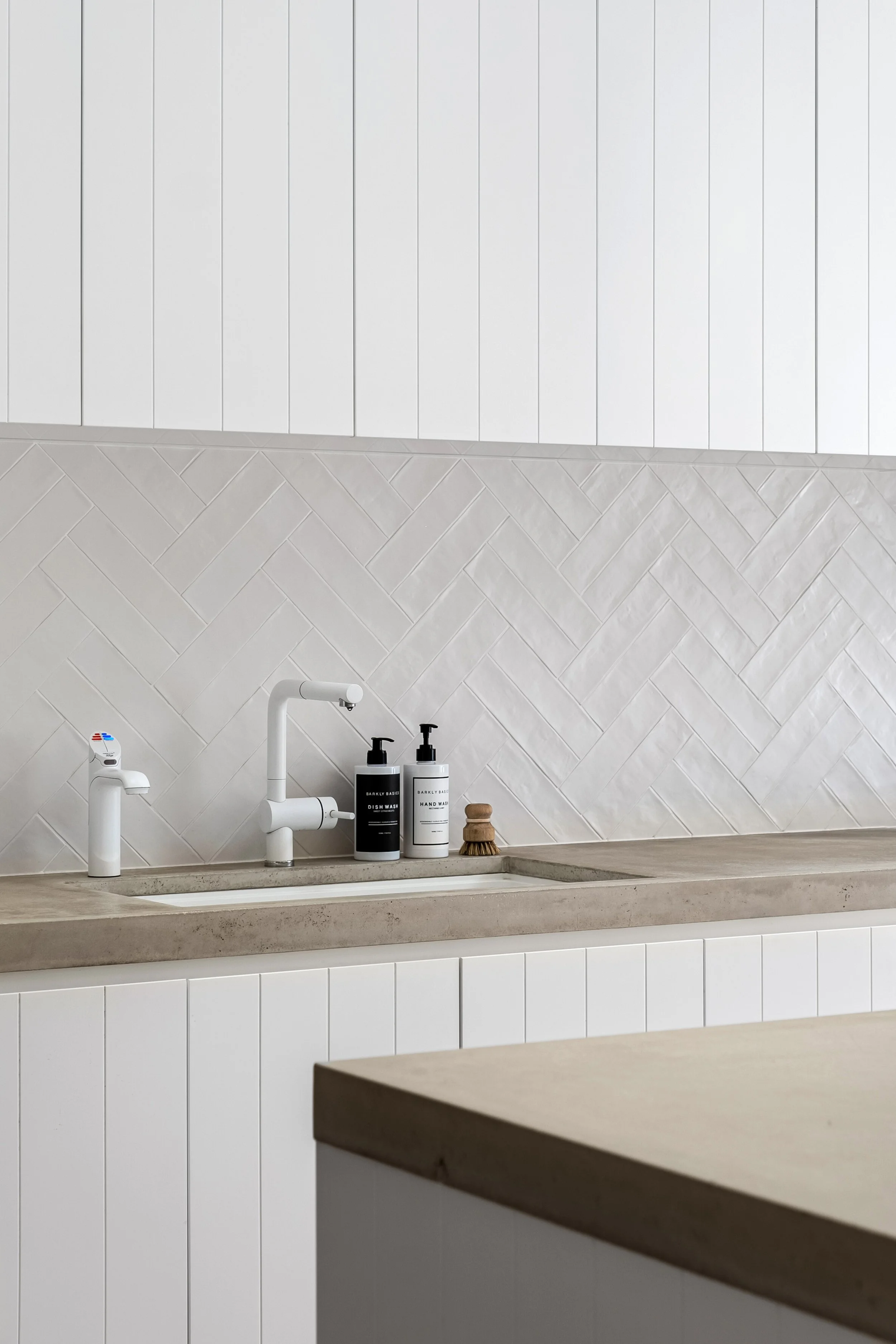 Minimalist kitchen sink area with white cabinets, a concrete countertop, and a white backsplash with a herringbone pattern. Contains soap dispensers, a brush, and a small container.