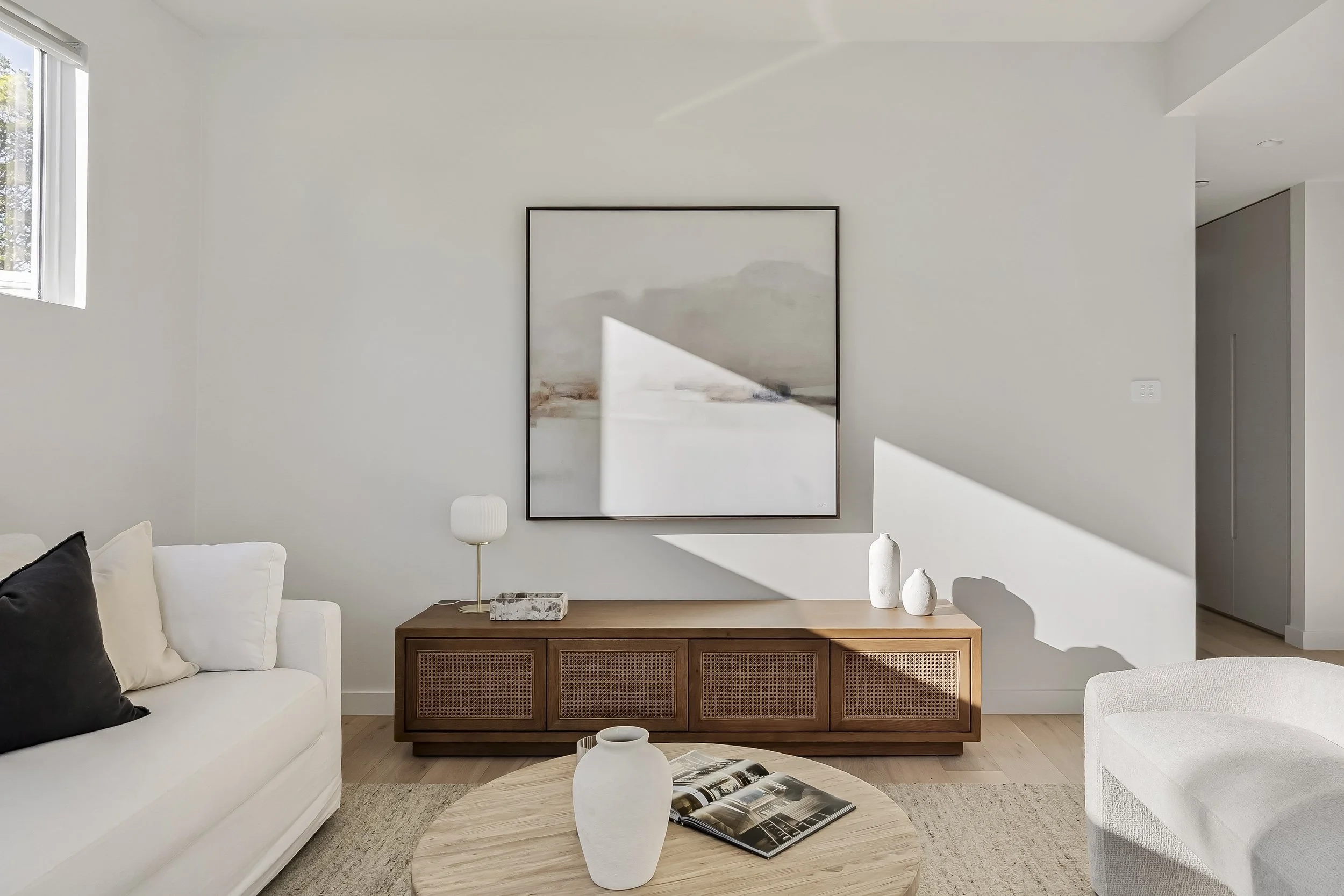 Minimalist living room with white sofa, black and white pillows, wooden sideboard, abstract art on the wall, white decorative vases, a round wooden coffee table with a book and a white vase, and sunlight creating shadows.