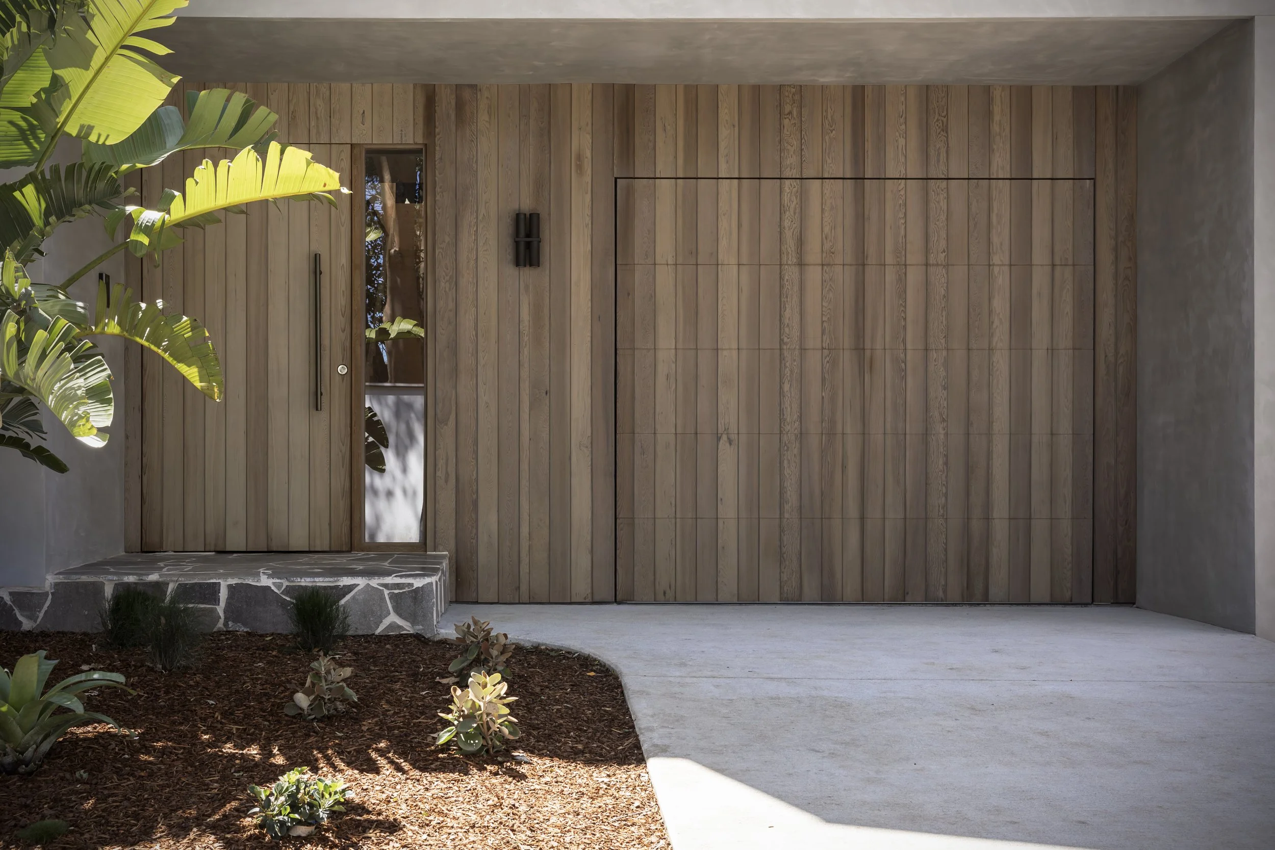Modern house entrance with a wooden garage door and a side door, surrounded by landscaping with plants and mulch.