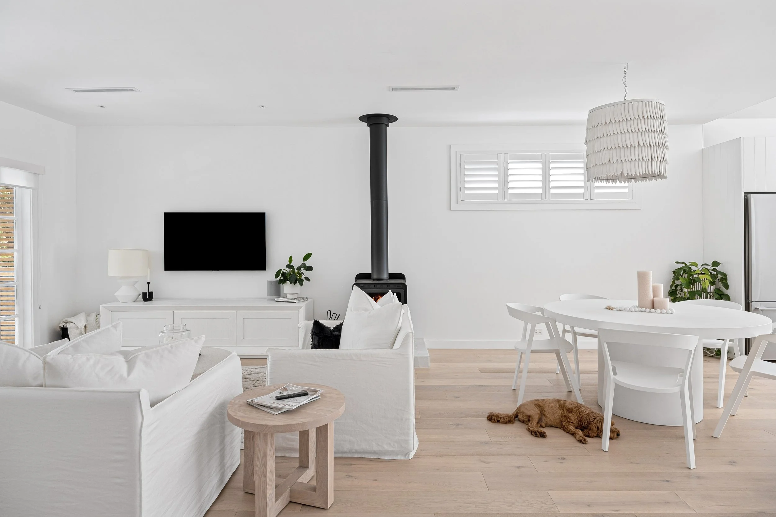 A modern, minimalist living room with white sofas, a round dining table, a black stove, and a sleeping brown dog on light wood flooring.