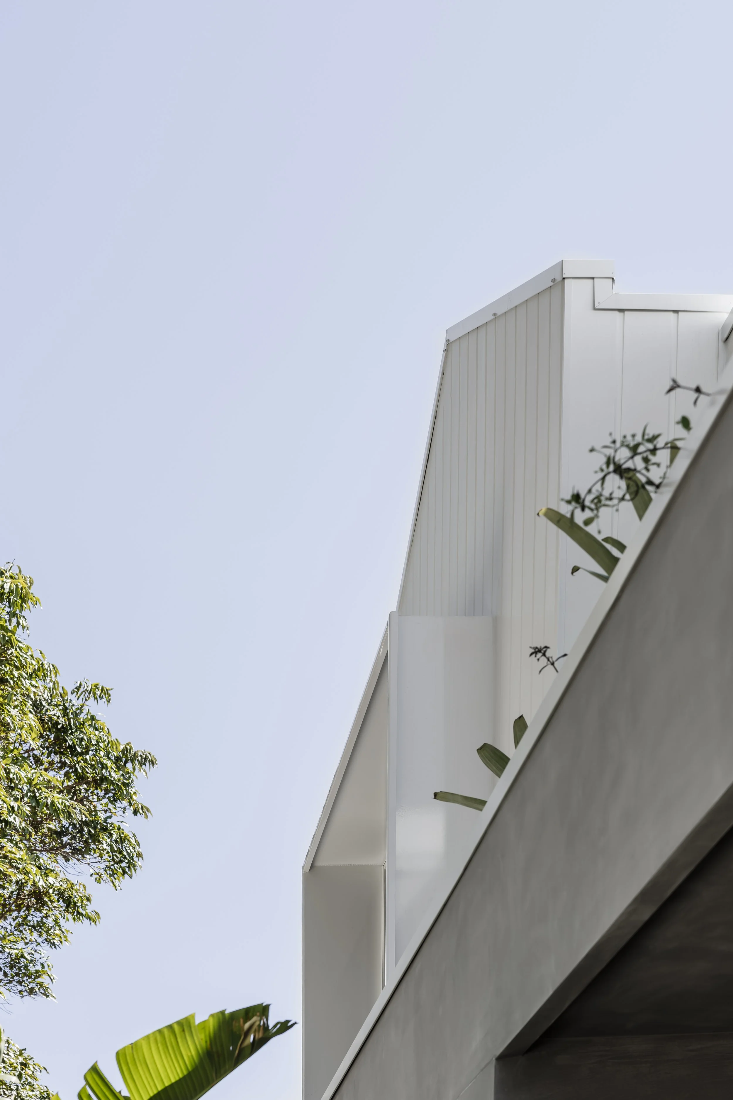 Close-up of a modern white building with a balcony, against a pale blue sky weather, with greenery and plants visible in the foreground