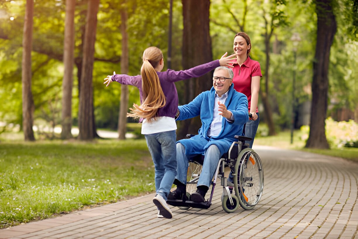 Family enjoying time outside; a young girl runs toward a smiling elderly man in a wheelchair, with a woman standing behind him, in a park setting with trees and a path.