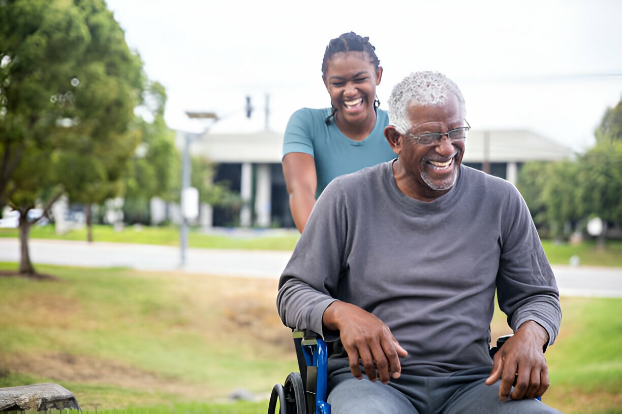 Smiling elderly man in wheelchair being pushed by a cheerful young woman outdoors.