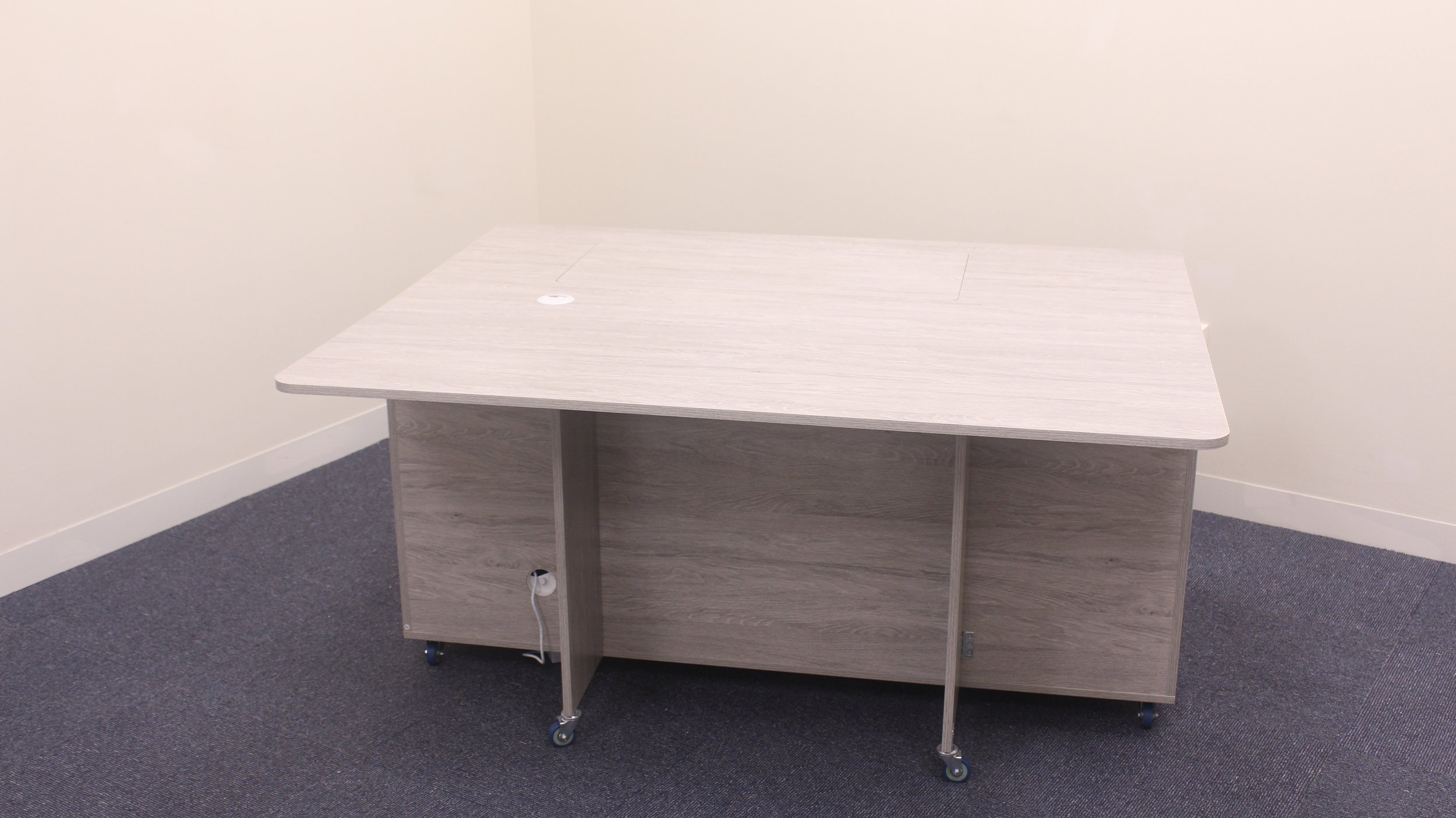 A light-colored wooden office table or desk with wheels, situated in a corner of a room with beige walls and dark gray carpeted floor.