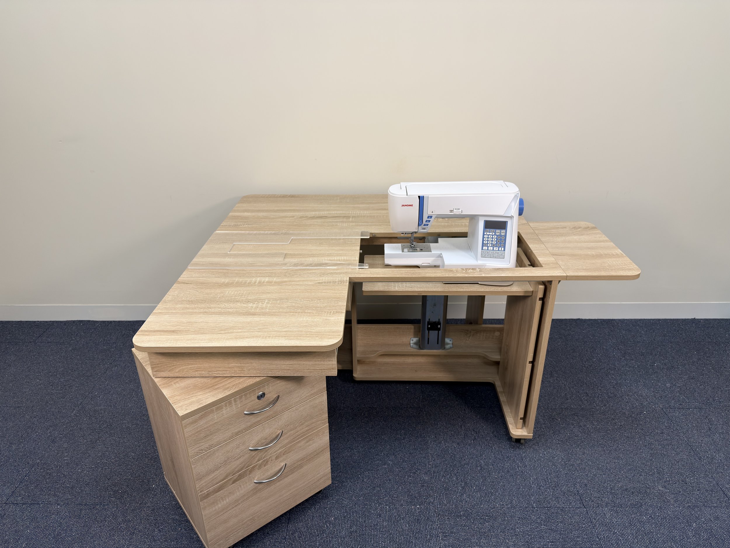 A wooden L-shaped desk with a white sewing machine placed on the middle section. There are three drawers on the left side of the desk and the right side has open shelves. The background is a plain beige wall and the floor is carpeted in dark blue.