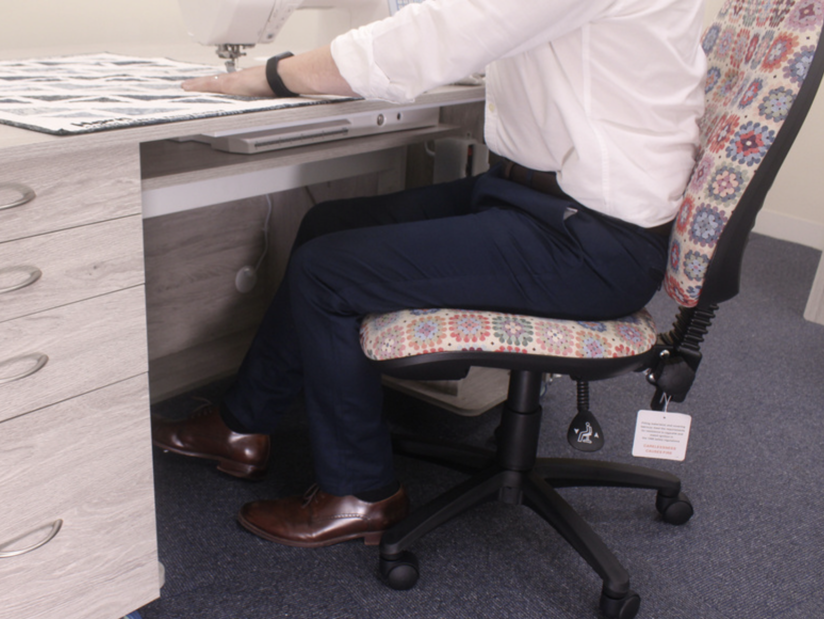 Person seated in an office chair at a desk, with their right hand reaching towards a sewing machine on the desk.